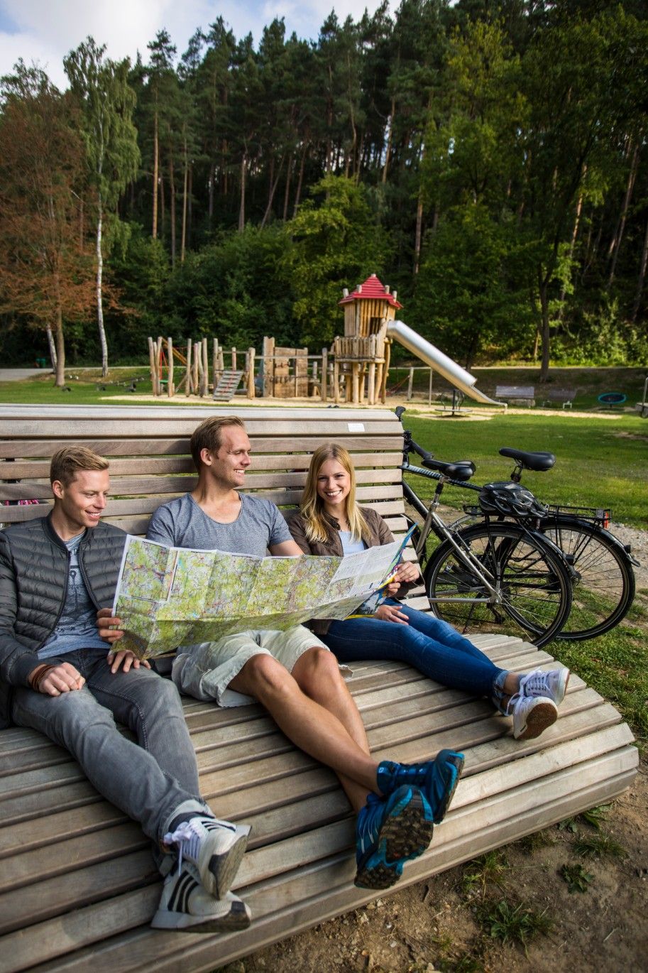 Drei Personen sitzen auf einer Bank im Freien und schauen auf eine Landkarte. Im Hintergrund ist ein Spielplatz und ein Wald zu sehen.