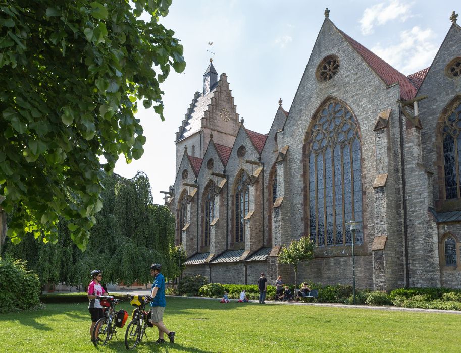 Zwei Radfahrer stehen vor einer Kirche im Grünen, umgeben von Bäumen und Menschen, die sich entspannen. Am dritten oder vierten Reisetag können Radreisende die Sehenswürdigkeiten im Osnabrücker Land ausmachen.