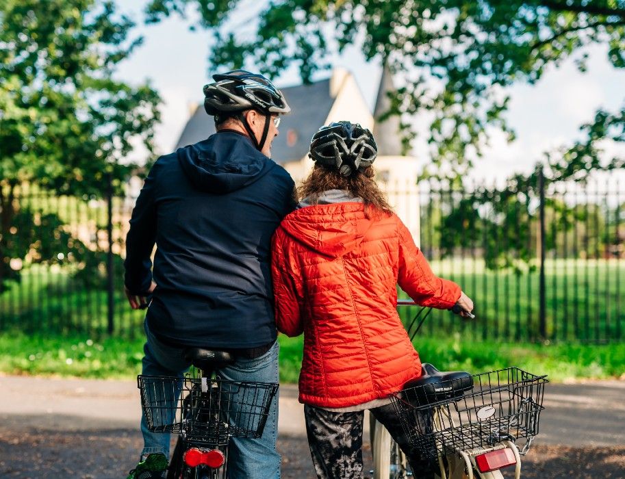 Zwei Radfahrer mit Helmen vor einem Zaun, im Hintergrund ein Gebäude.