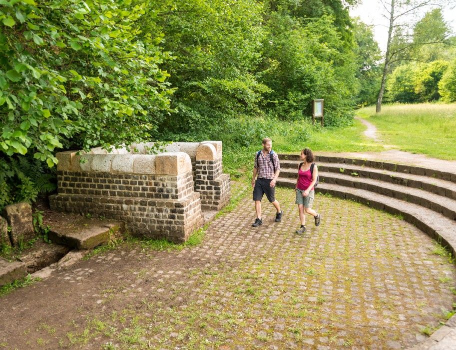Zwei Personen wandern auf einem gepflasterten Weg neben einer historischen Brunnenanlage im Grünen.