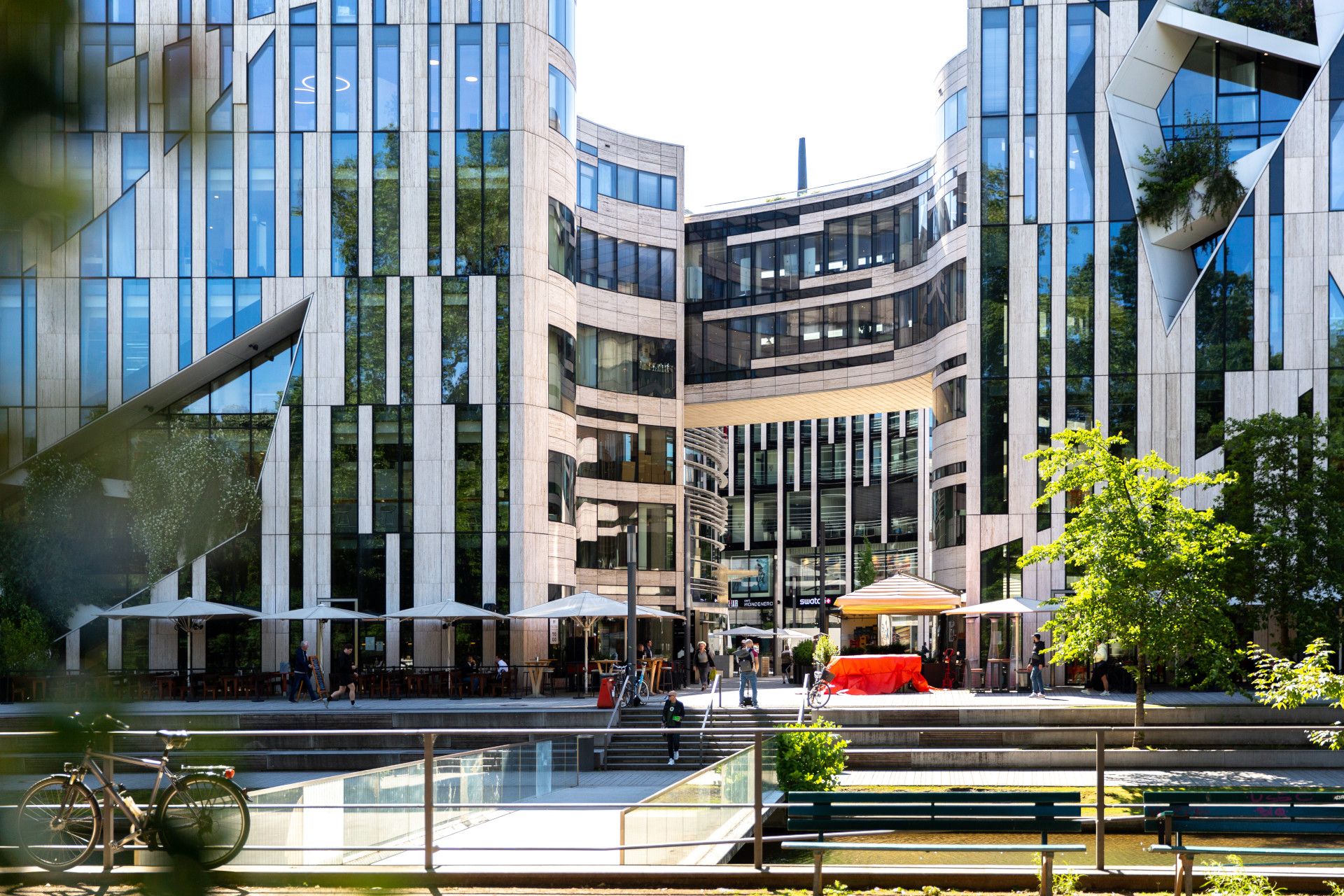 Der Kö-Bogen in Düsseldorf zeigt moderne Architektur mit Glasfassaden und urbanem Flair. Menschen flanieren, während Fahrräder und Bäume das Bild beleben.