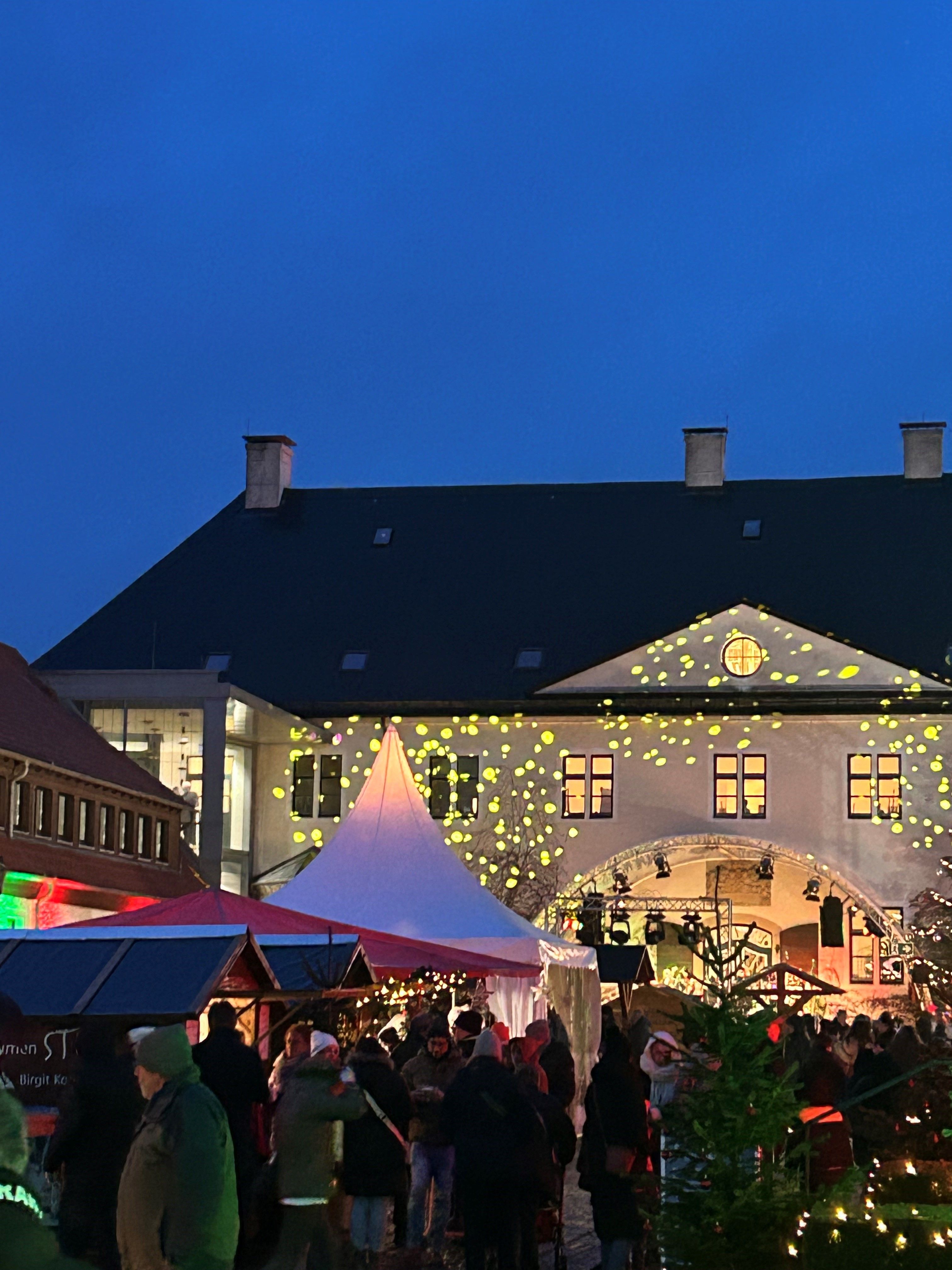 Weihnachtsmarkt vor Schloss Benkhausen bei Nacht. Menschen schlendern zwischen festlich beleuchteten Ständen. Lichterprojektionen schmücken die Schlossfassade.