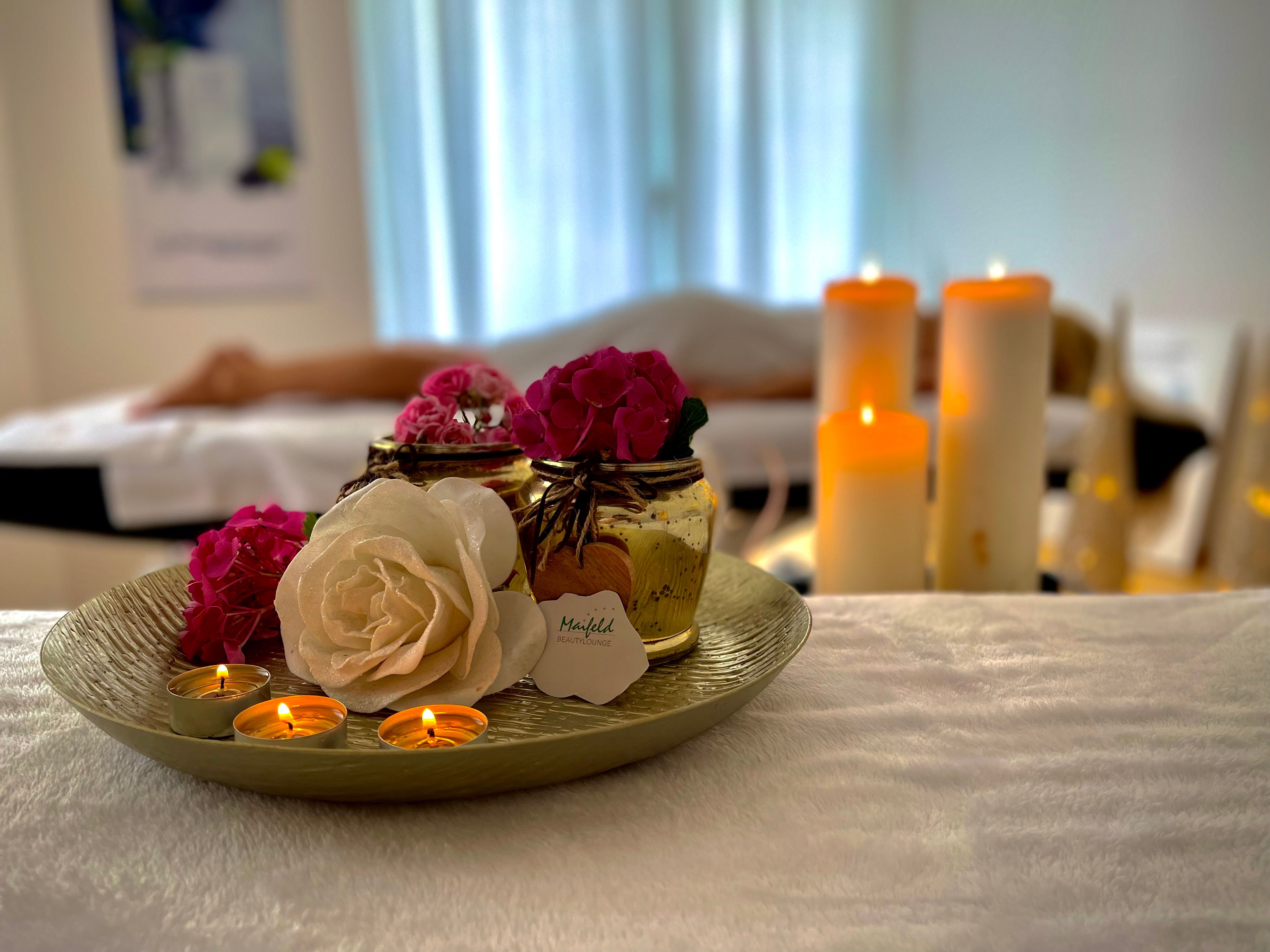 Ein entspannender Raum in einem Beautysalon mit Kerzen, Blumen und einer liegenden Person im Hintergrund.
