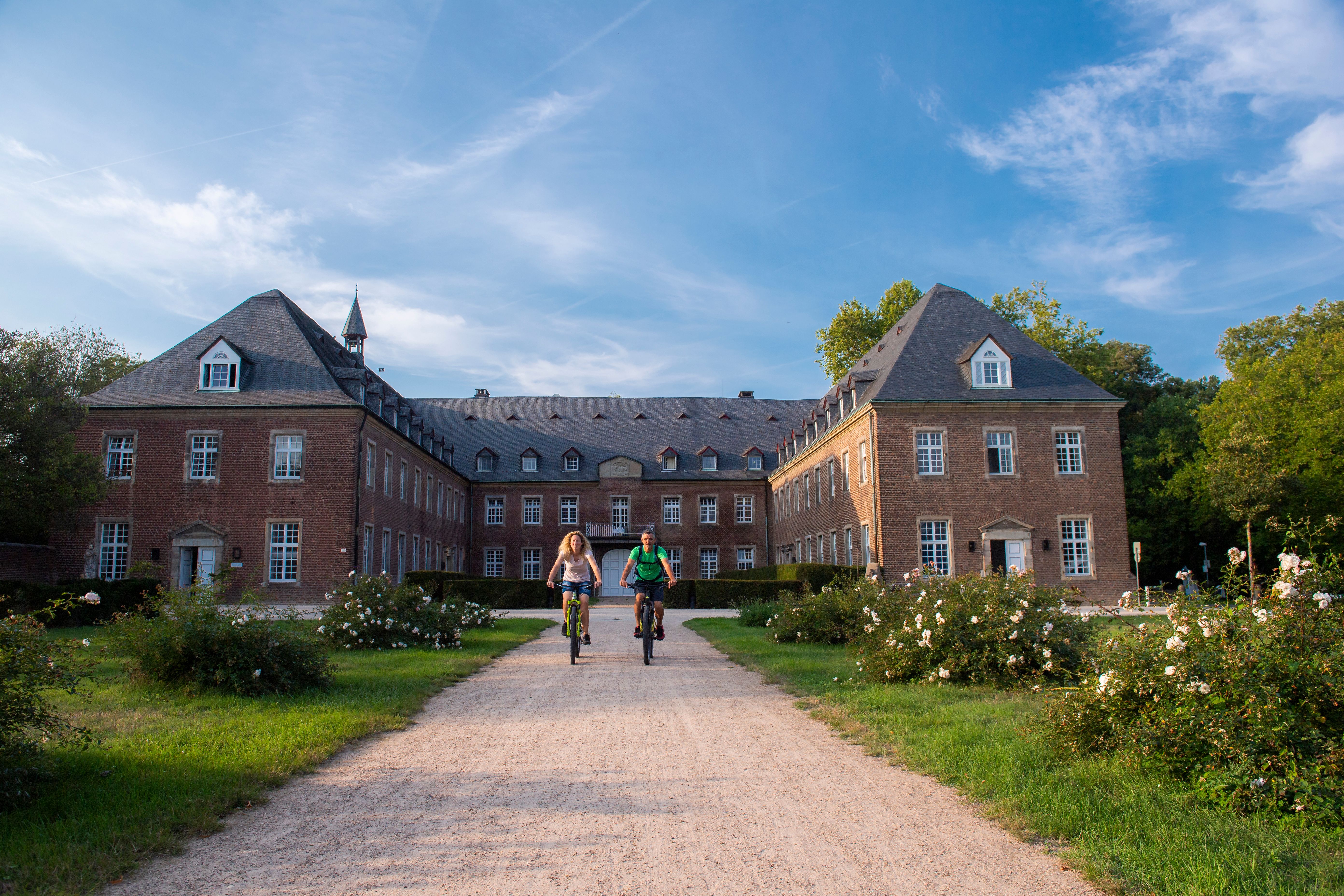 Zwei Personen fahren mit Fahrrädern auf einem Weg vor dem Kloster Langwaden mit Ziegeldach und vielen Fenstern.