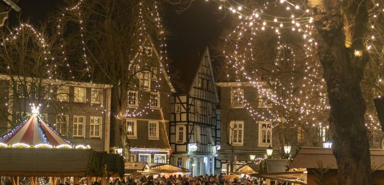 Nächtlicher Weihnachtsmarkt mit Lichterketten, Fachwerkhäusern und Menschenmenge.