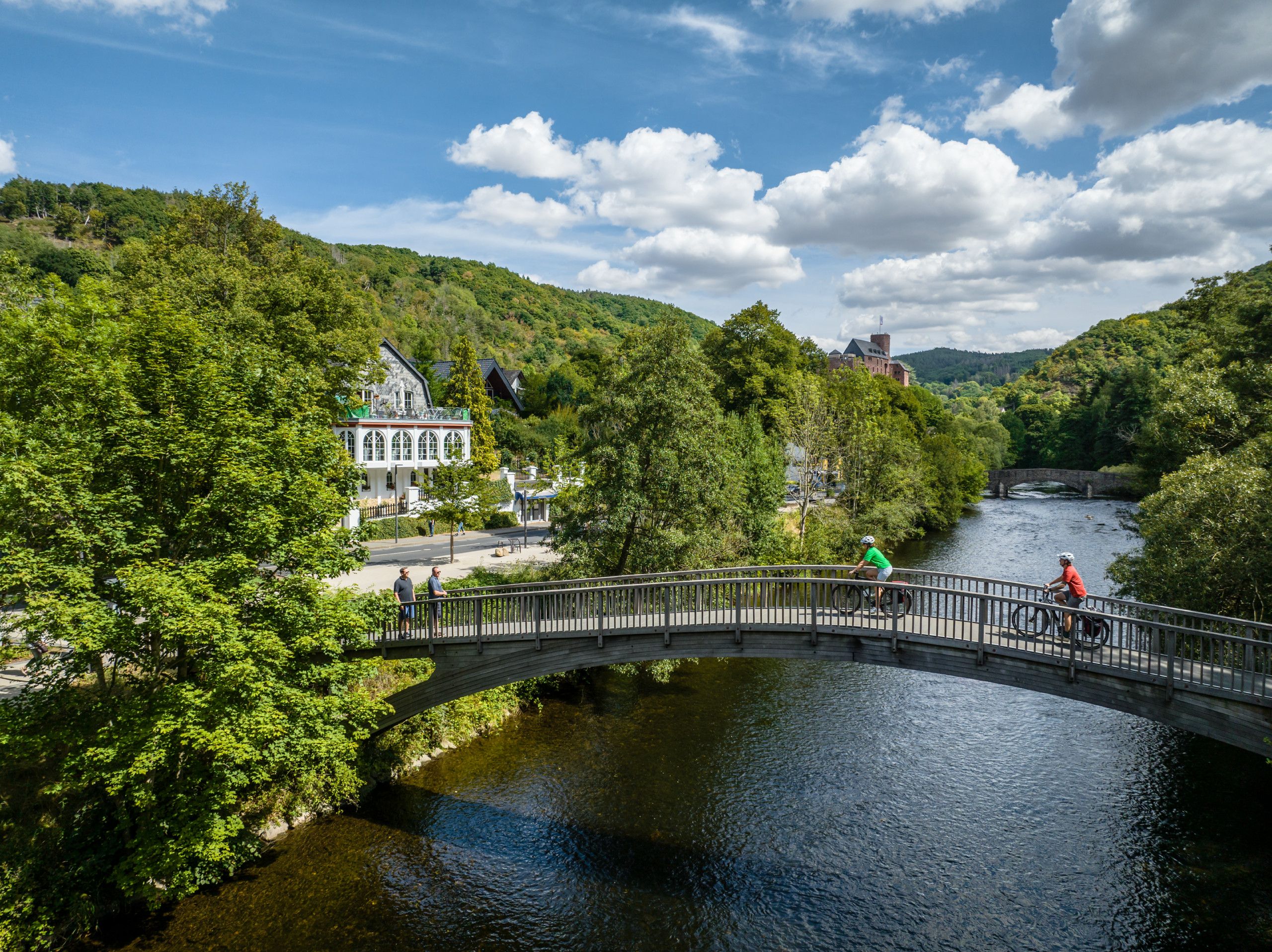 Brücke über Fluss in Heimbach mit Radfahrern und grüner Landschaft.
