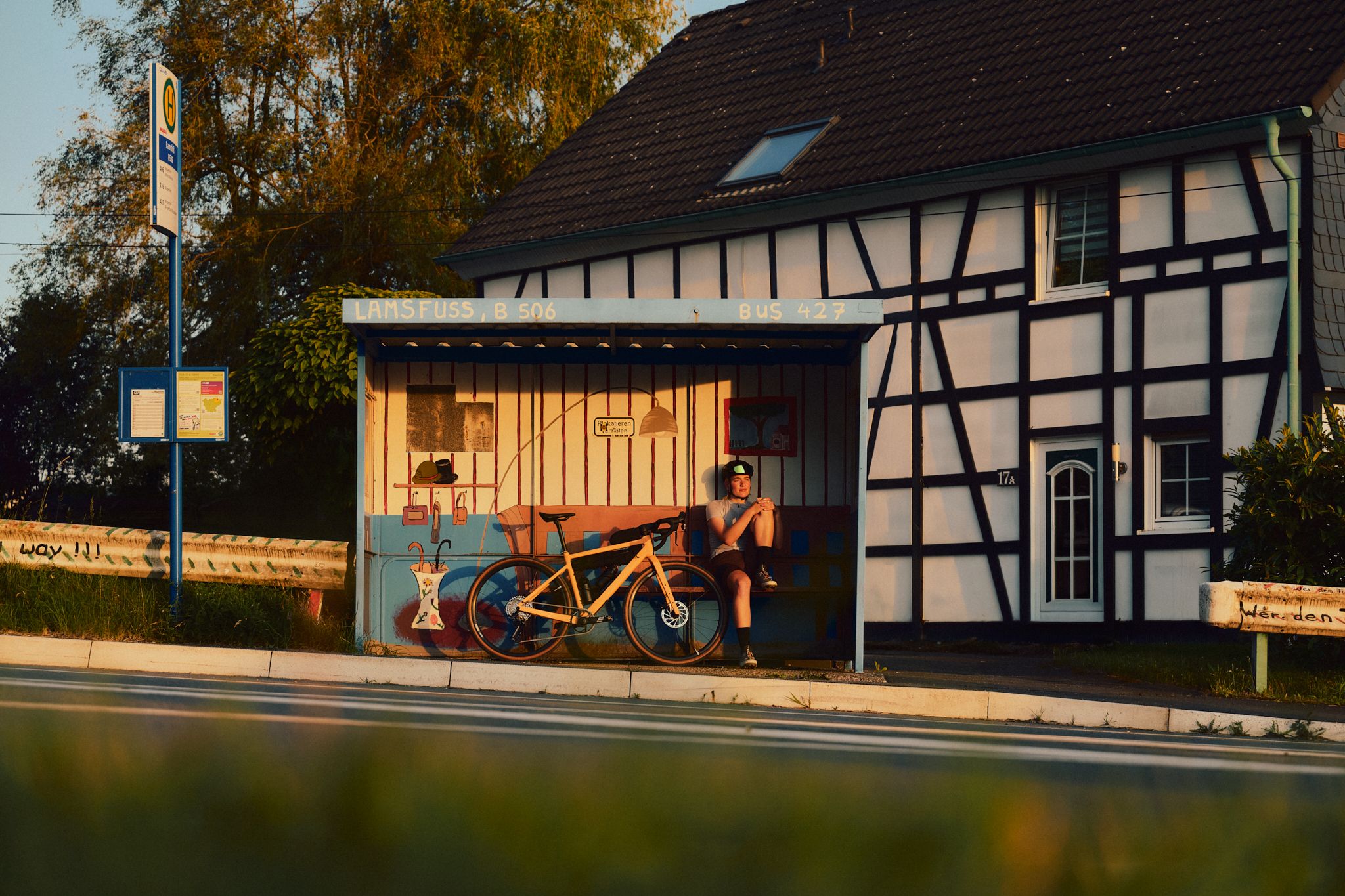 Person sitzt mit Fahrrad an einer Bushaltestelle vor einem Fachwerkhaus.
