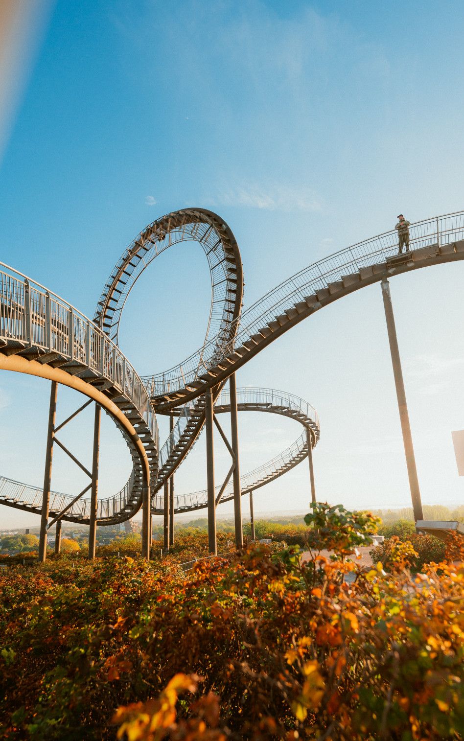 Die begehbare Skulptur Tiger & Turtle in Duisburg windet sich in einer Achterbahnform unter einem farbenfrohen Abendhimmel.Tiger & Turtle Skulptur in Duisburg bei Sonnenuntergang.