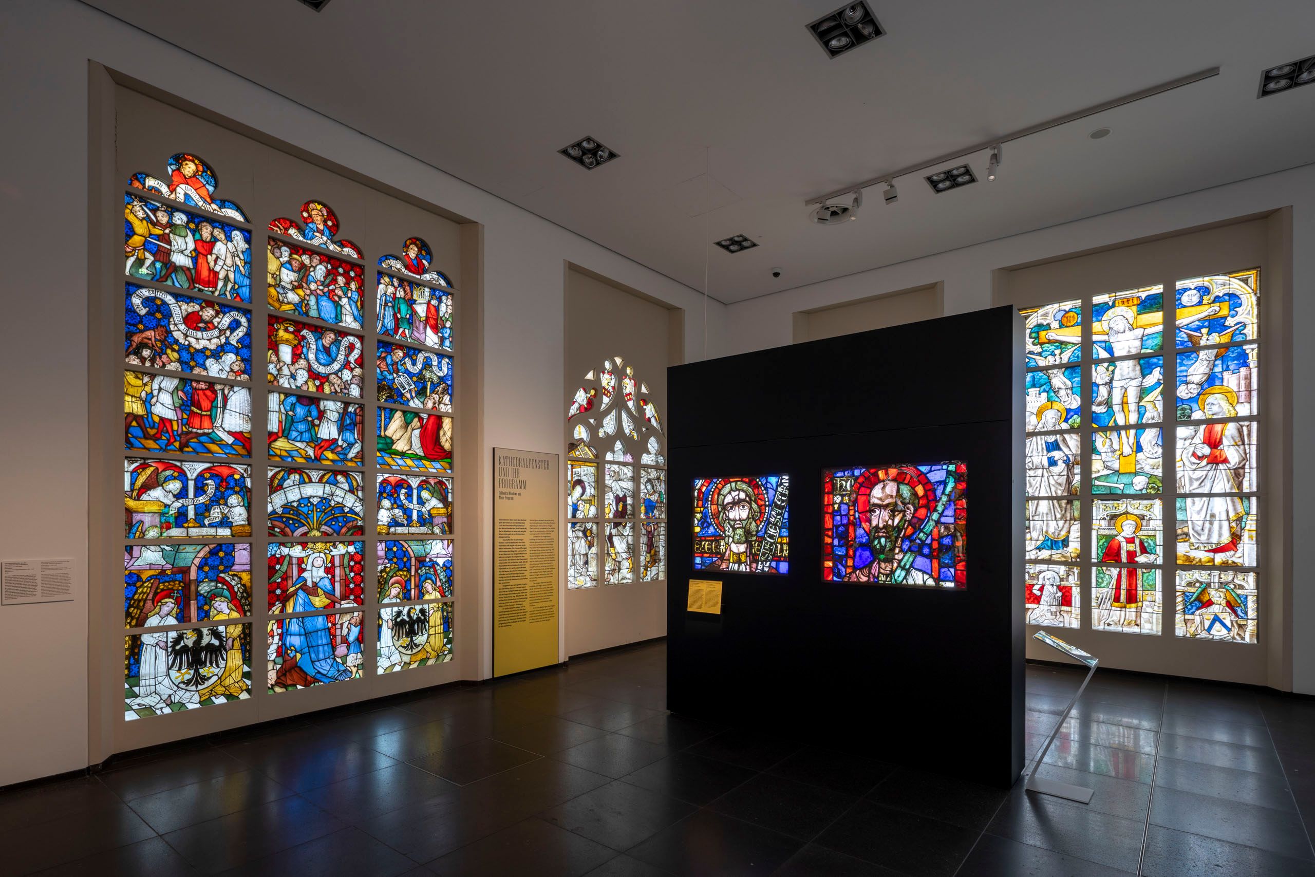 Buntglasfenster im Museum Schnütgen in Köln, die religiöse Szenen darstellen. Die Fenster sind kunstvoll gestaltet und farbenfroh.