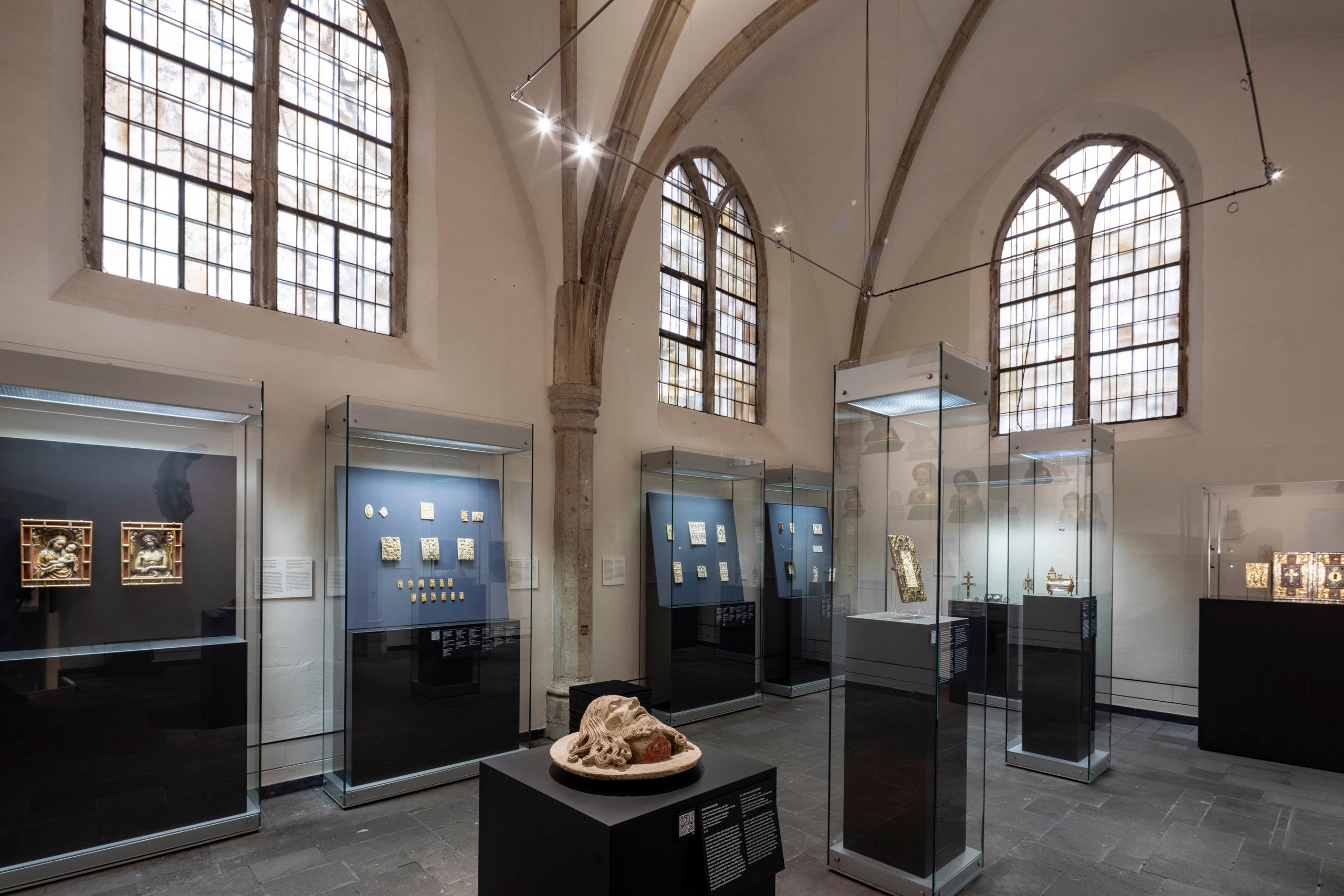 Ausstellungsraum im Museum Schnütgen in Köln mit mittelalterlichen Kunstwerken in Glasvitrinen und gotischen Fenstern im Hintergrund.