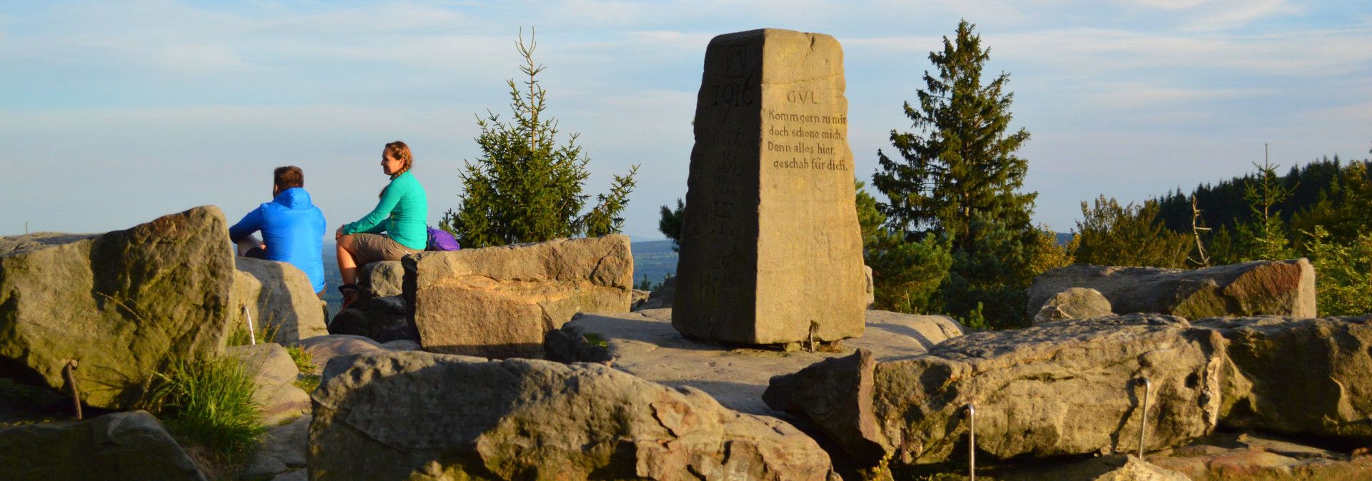 Zwei Personen sitzen auf Felsen am Lippischen Velmerstot mit einem Gedenkstein im Vordergrund.