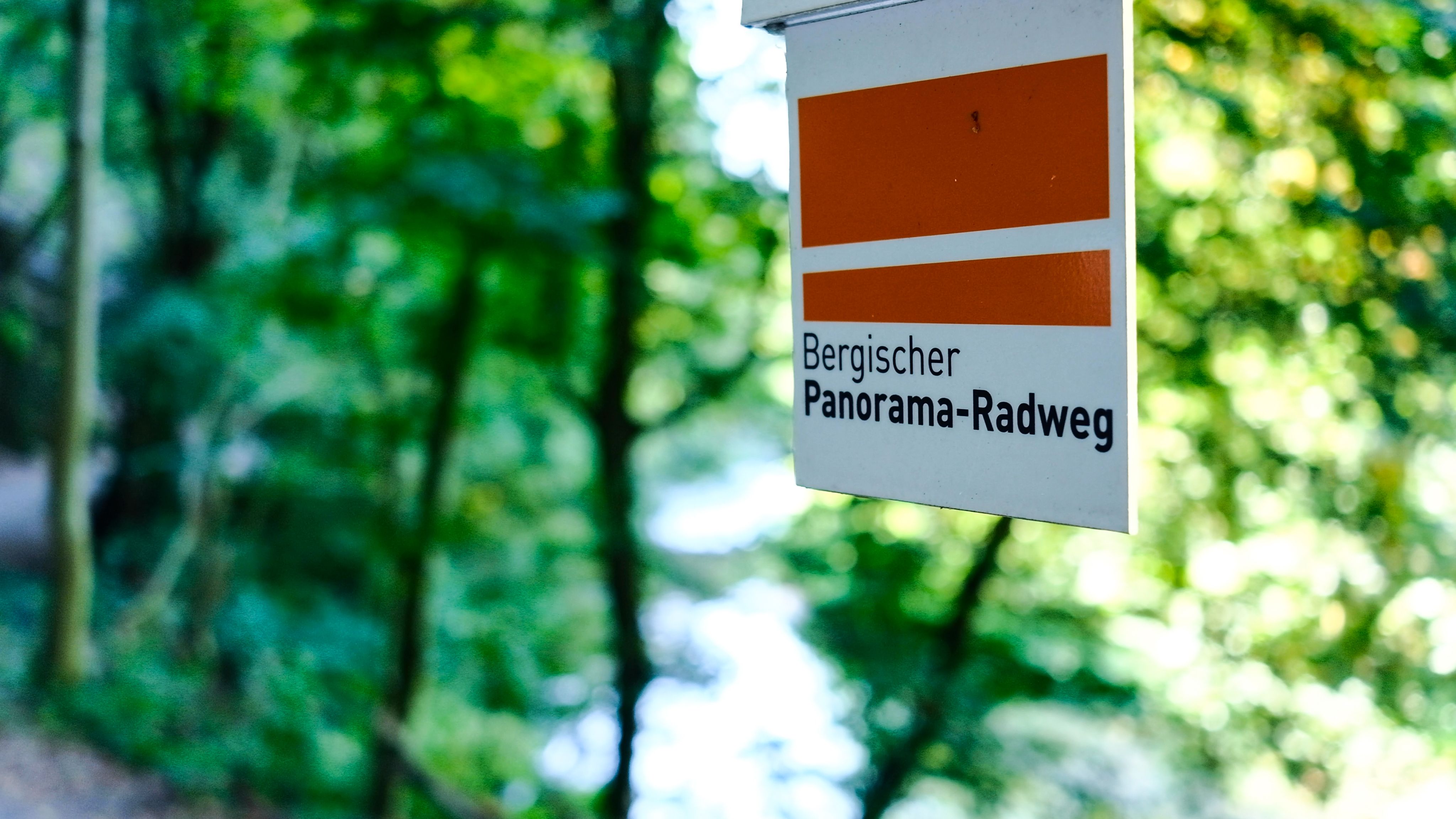 Ein Schild mit der Aufschrift 'Bergischer Panorama-Radweg' vor einem verschwommenen, grünen Waldhintergrund.