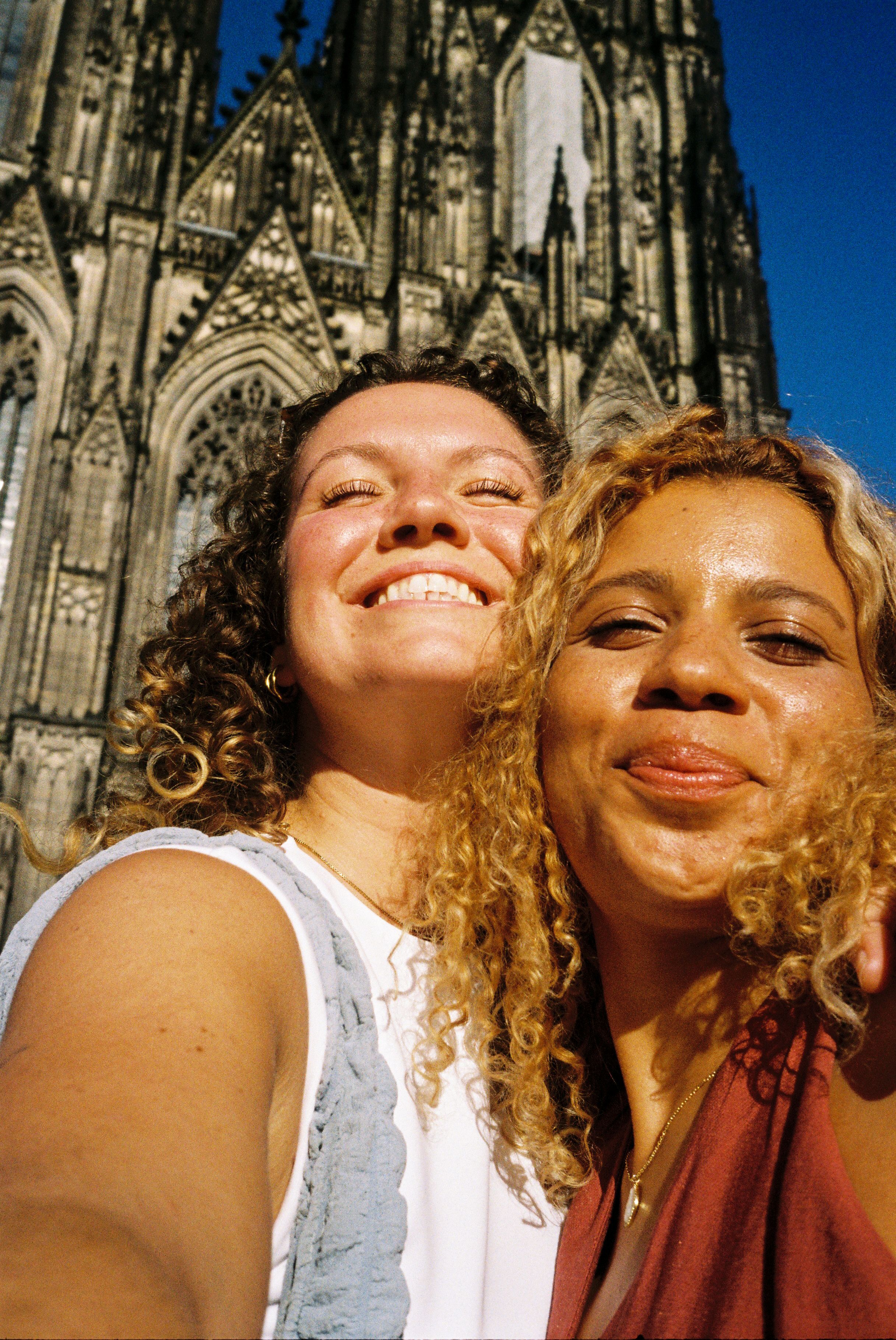 Selfie von zwei Frauen im Sonnenschein lächelnd vor dem Kölner Dom.