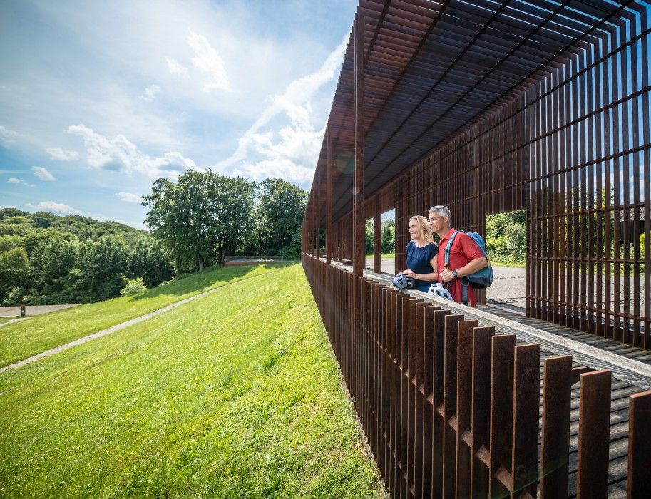 Zwei Personen betrachten von einer rostfarbenen Metallstruktur aus eine grüne Landschaft. Der Himmel ist blau mit wenigen Wolken.
