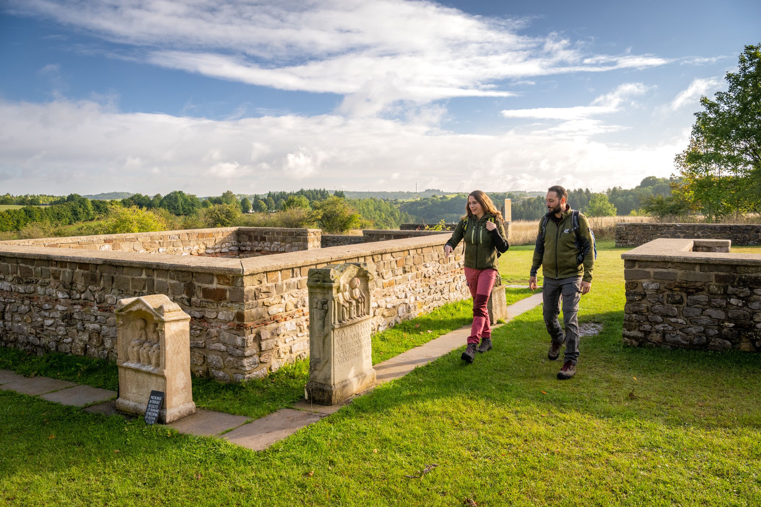 Zwei Wanderer spazieren am Matronenheiligtum Görresburg vorbei. Im Hintergrund ist eine grüne Landschaft zu sehen.