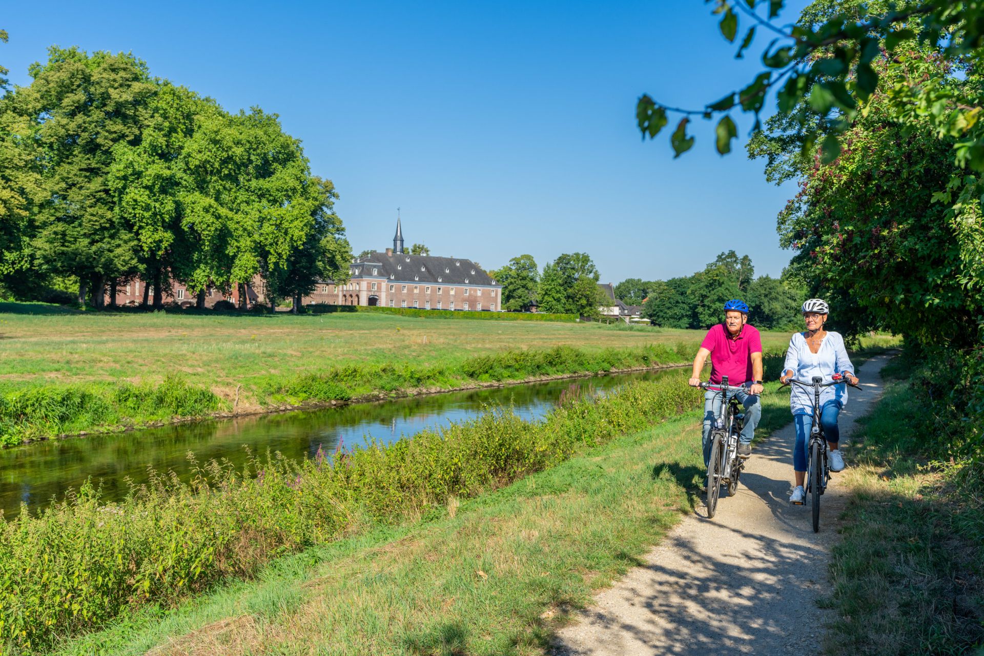 Zwei Radfahrer auf einem Weg entlang eines Flusses mit einem Gebäude im Hintergrund.