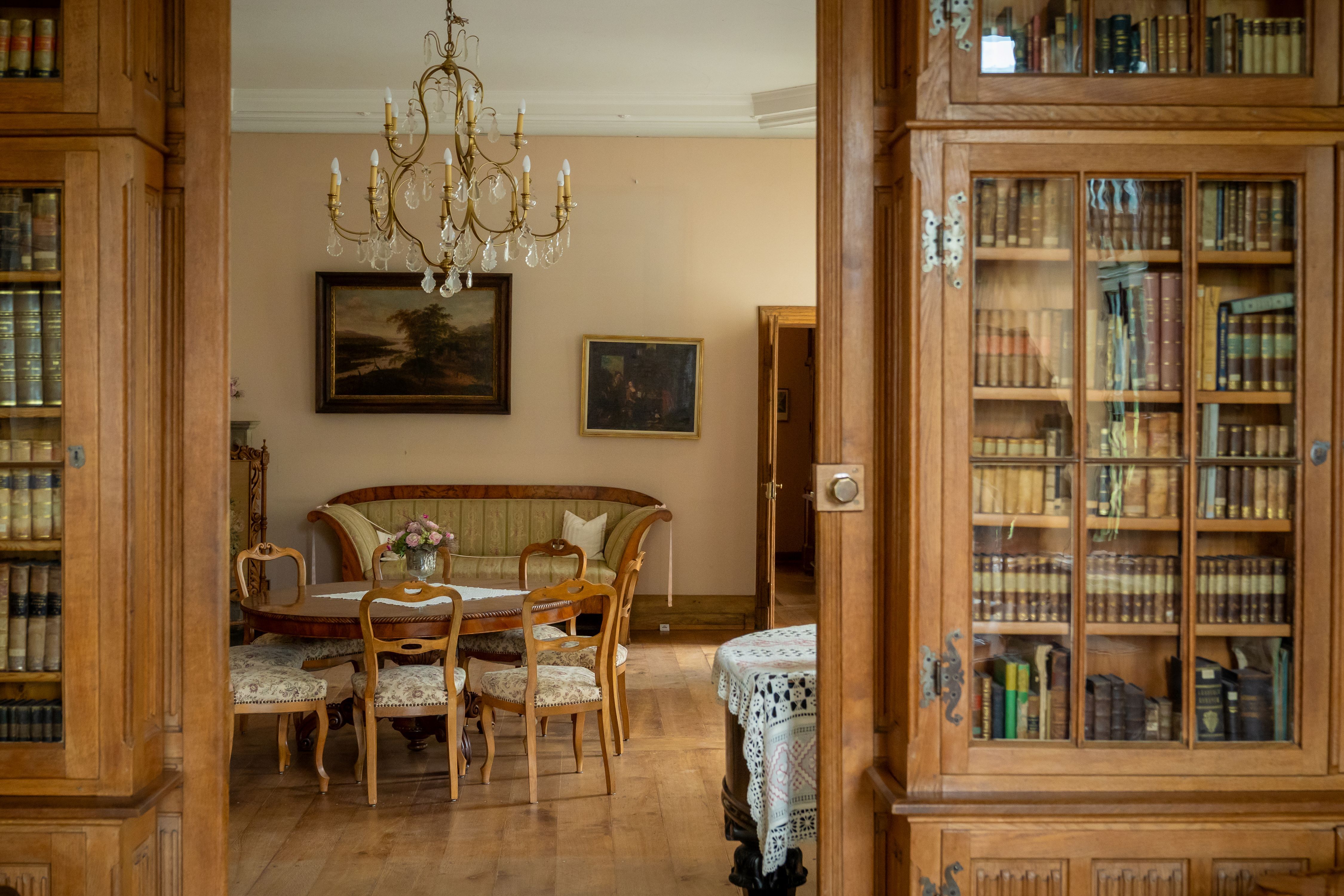 Blick vorbei an Bücherschränken in einen Salon mit Sitzgruppe und Bildern an der Wand.
