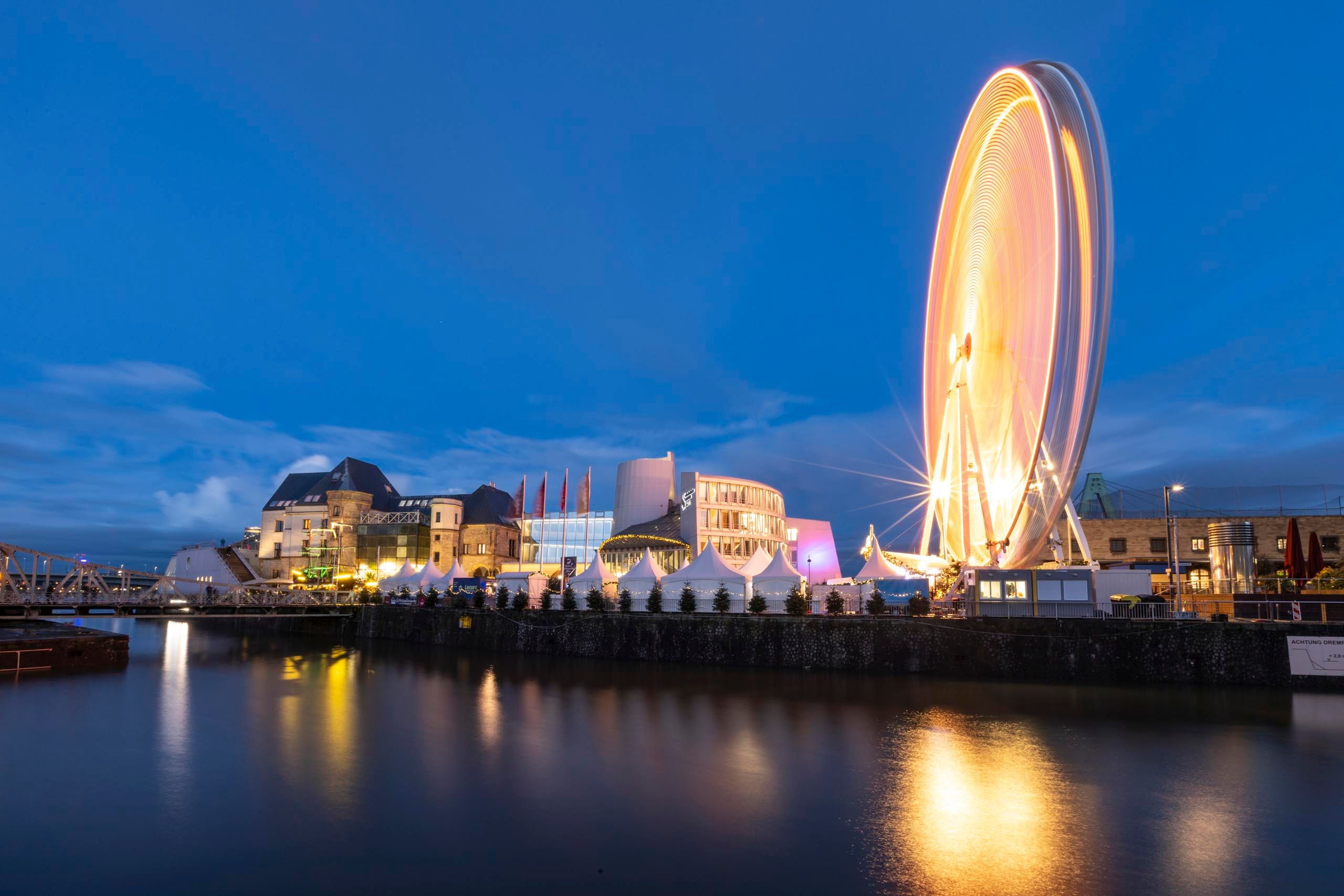 Abendaufnahme des Kölner Hafen-Weihnachtsmarkts mit beleuchtetem Riesenrad und Gebäuden am Wasser.