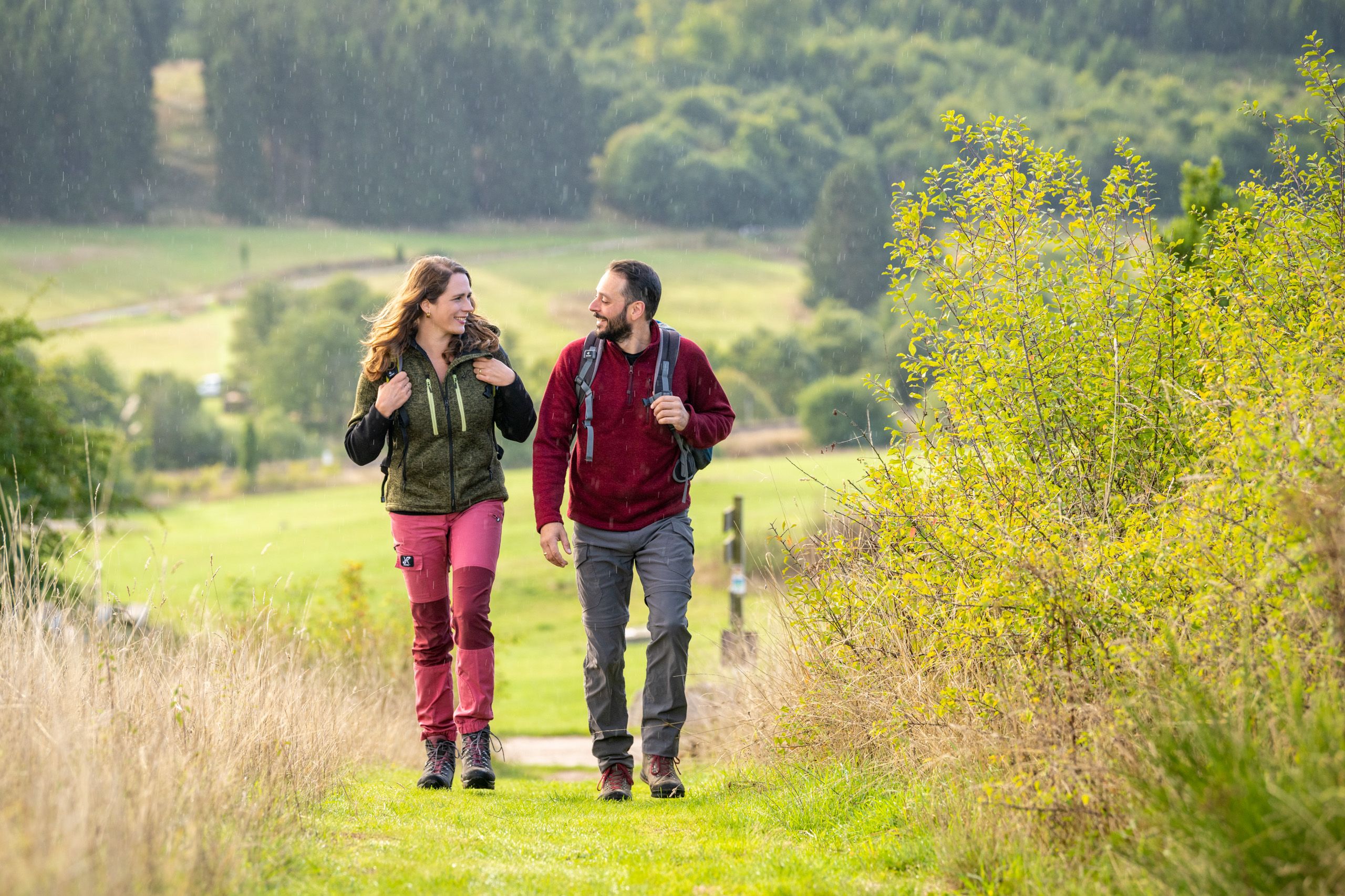 Zwei Personen wandern mit Rucksäcken durch eine grüne Landschaft und unterhalten sich.