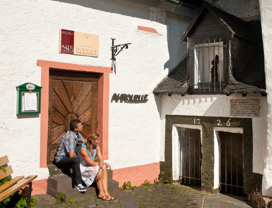 Zwei Personen sitzen vor der Ahrquelle in Blankenheim, Eifel. Die Fassade ist weiß mit einem Holztor und Schildern. Baujahr 1726 ist sichtbar.