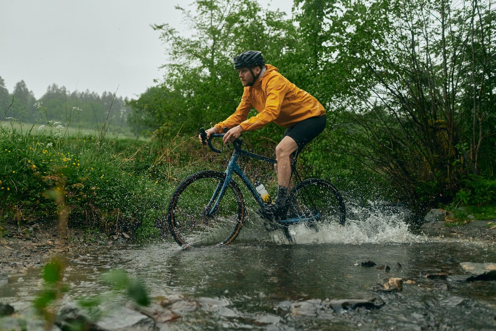 Ein Radfahrer in gelber Jacke fährt durch einen flachen Bach in einer grünen Landschaft.