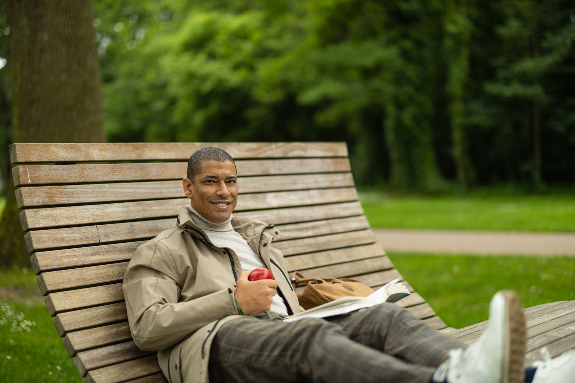 Ein Mann sitzt entspannt auf einer Holzbank im Park und hält einen roten Apfel in der Hand.