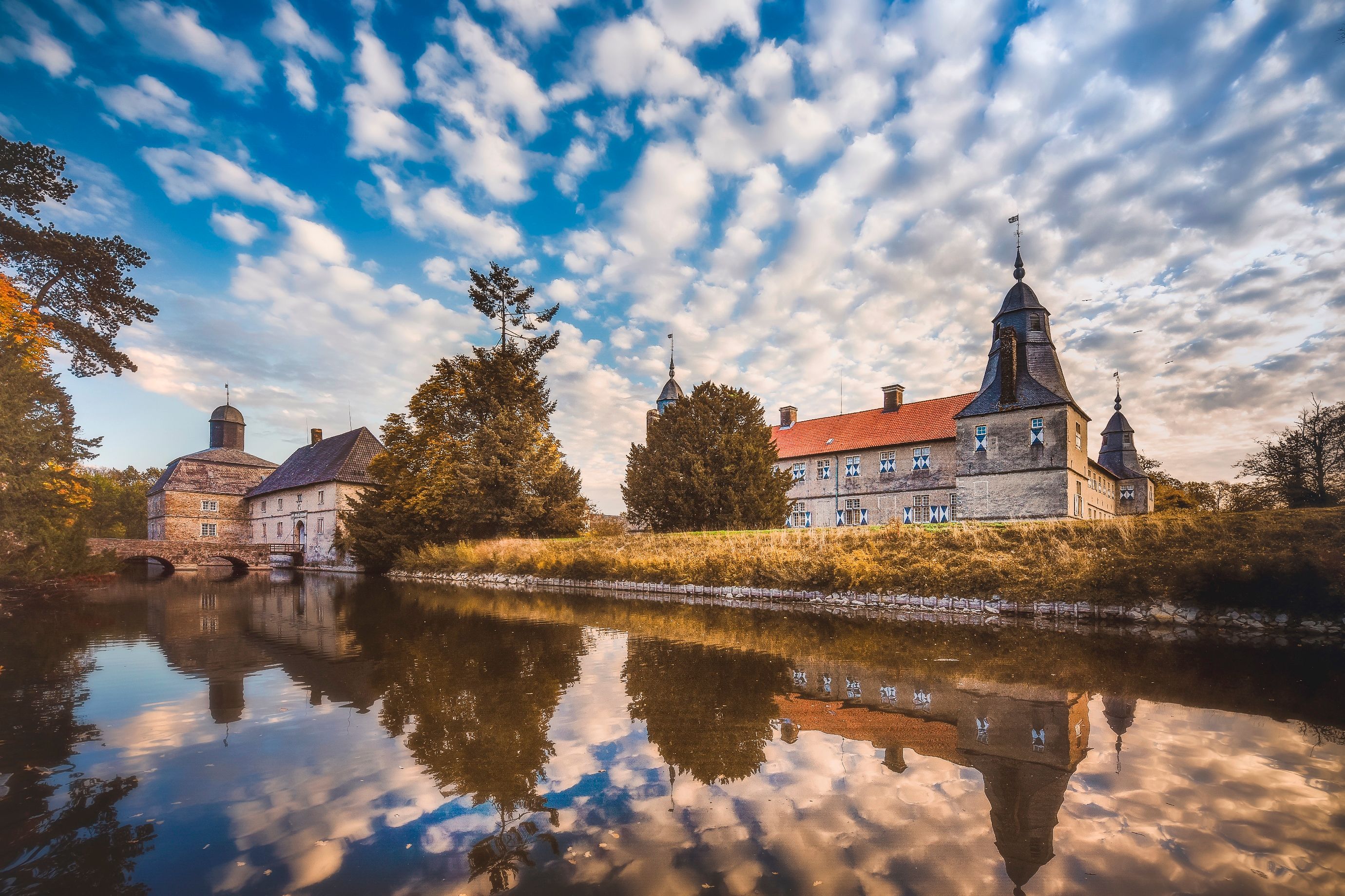 Schloss Westerwinkel in Herbern, umgeben von einem Wassergraben. Die Gebäude spiegeln sich im ruhigen Wasser, der Himmel ist mit Wolken bedeckt.