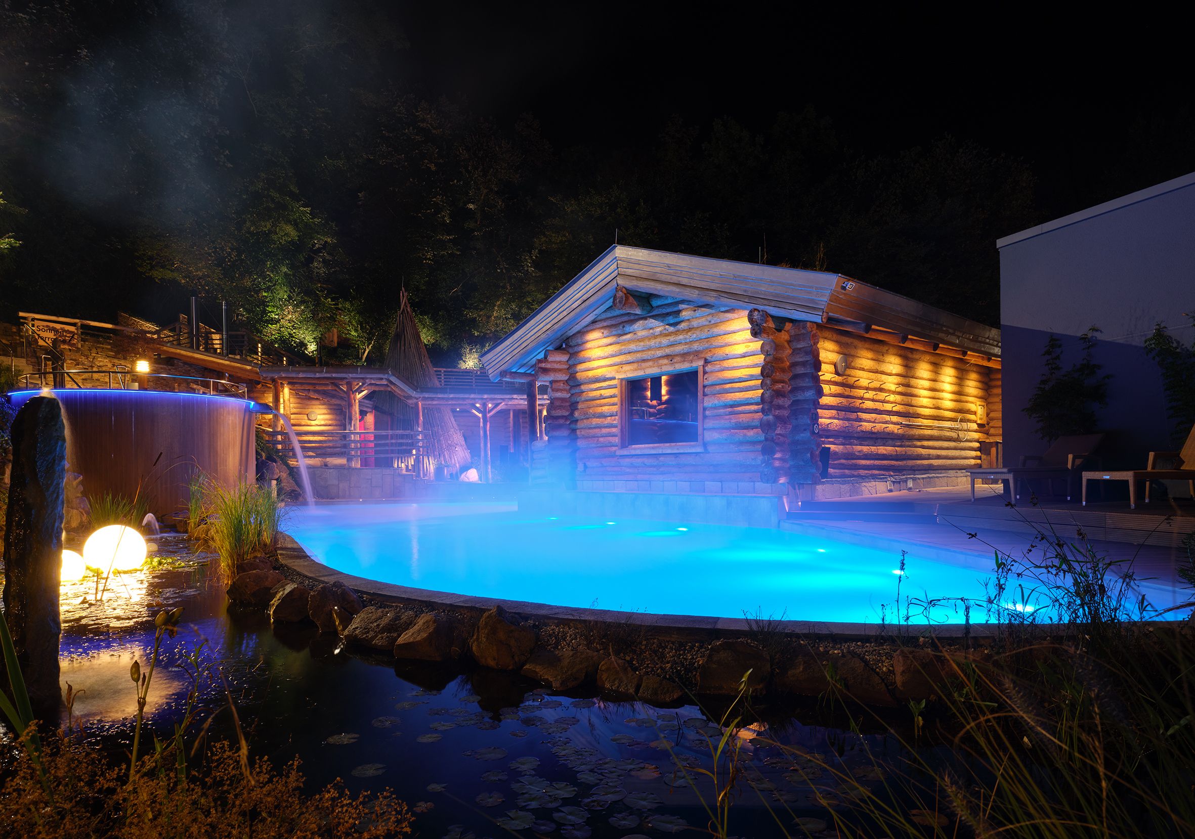 Beleuchtete Therme mit Holzgebäude und Pool bei Nacht.