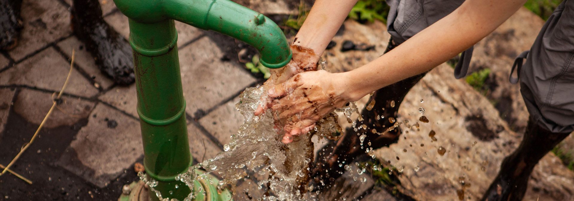 Person wäscht schlammige Hände an einer grünen Wasserpumpe.