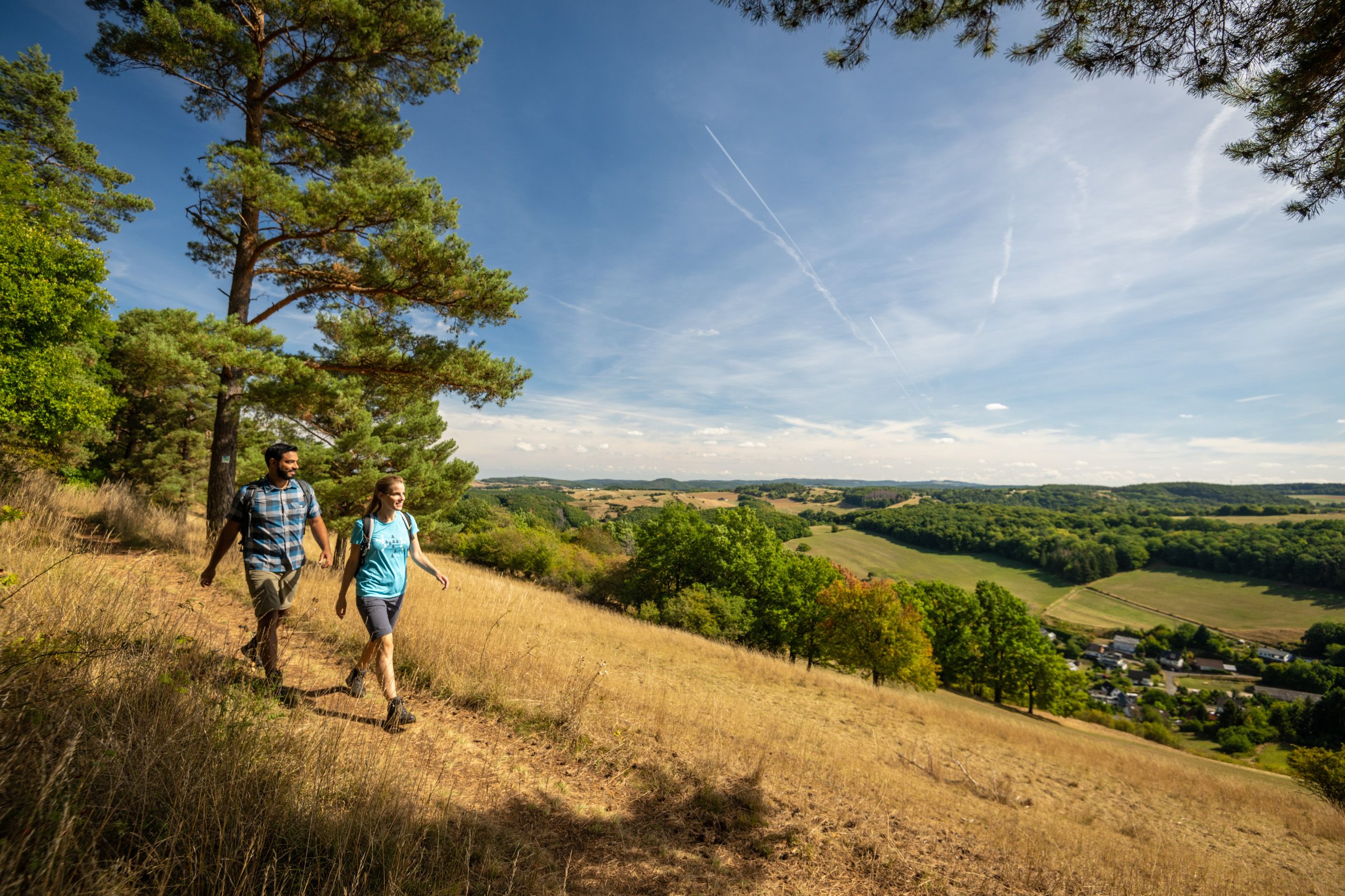 Zwei Wanderer auf einem Pfad mit Blick auf eine weite Landschaft und blauen Himmel.