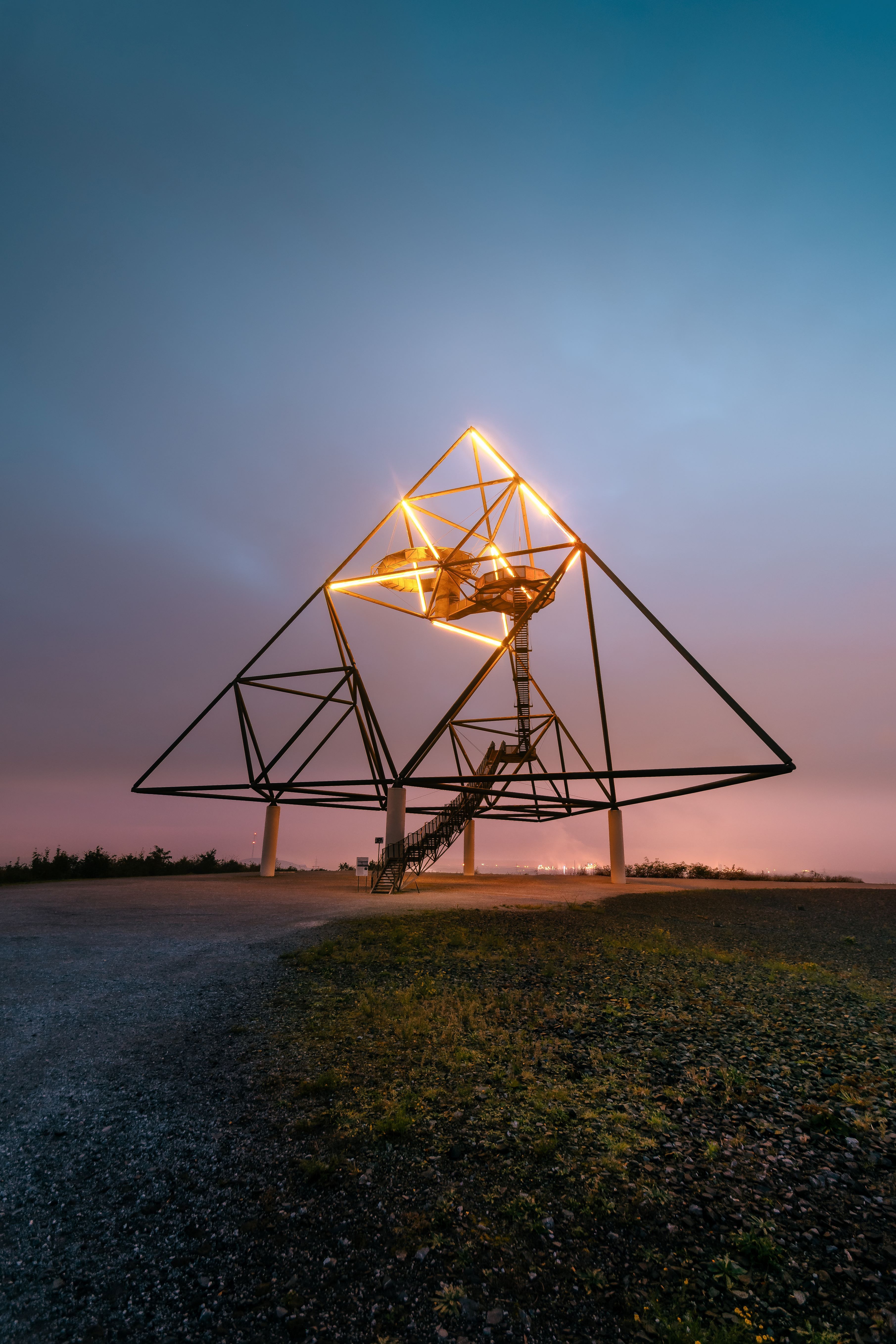 Auf einer Halde bei Dämmerung steht die Skulptur eines Tetraeders mit beleuchteter Spitze