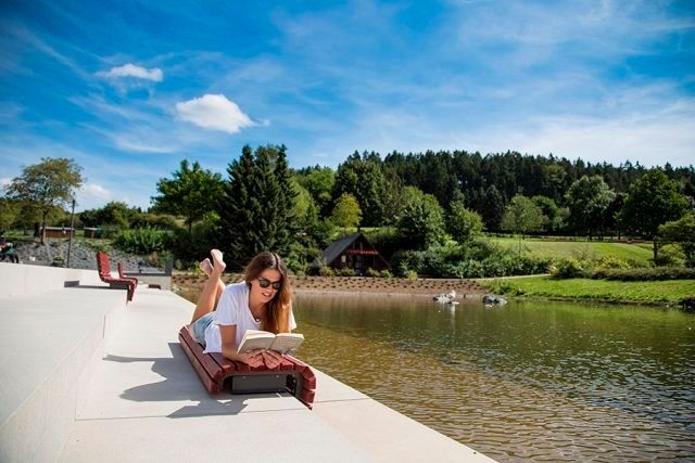 Eine Frau liegt auf einer Bank am Teich im Kurpark und liest ein Buch. Im Hintergrund sind Bäume und ein klarer Himmel zu sehen.