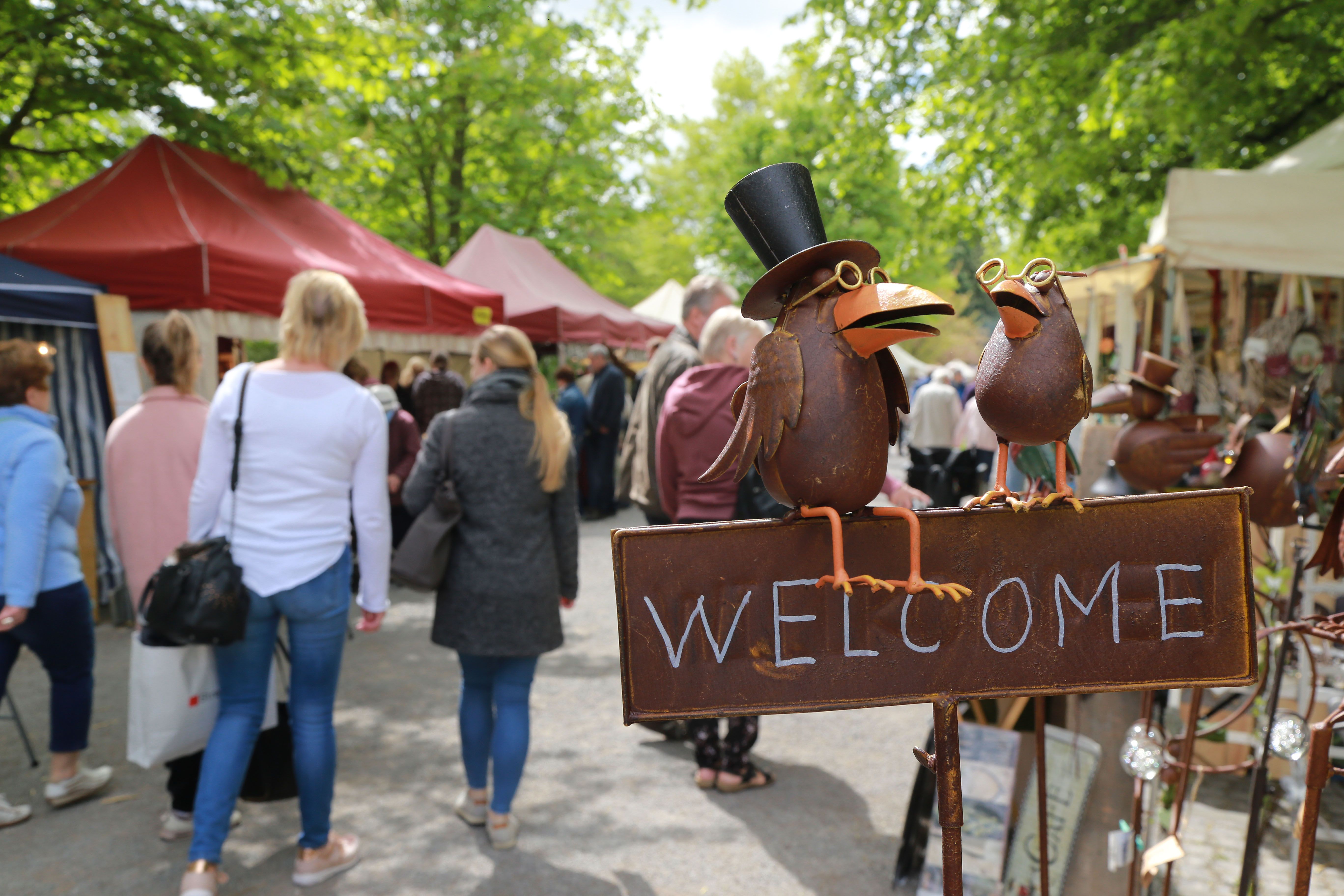 Metallvögel mit Zylinder und Brille auf einem Schild mit der Aufschrift 'Welcome' auf dem Handwerker- und Bauernmarkt in Bad Sassendorf mit Menschen und Ständen im Hintergrund.