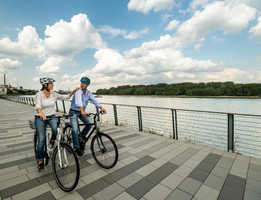 Zwei Personen fahren auf Fahrrädern entlang des Rheins auf einem gepflasterten Weg.