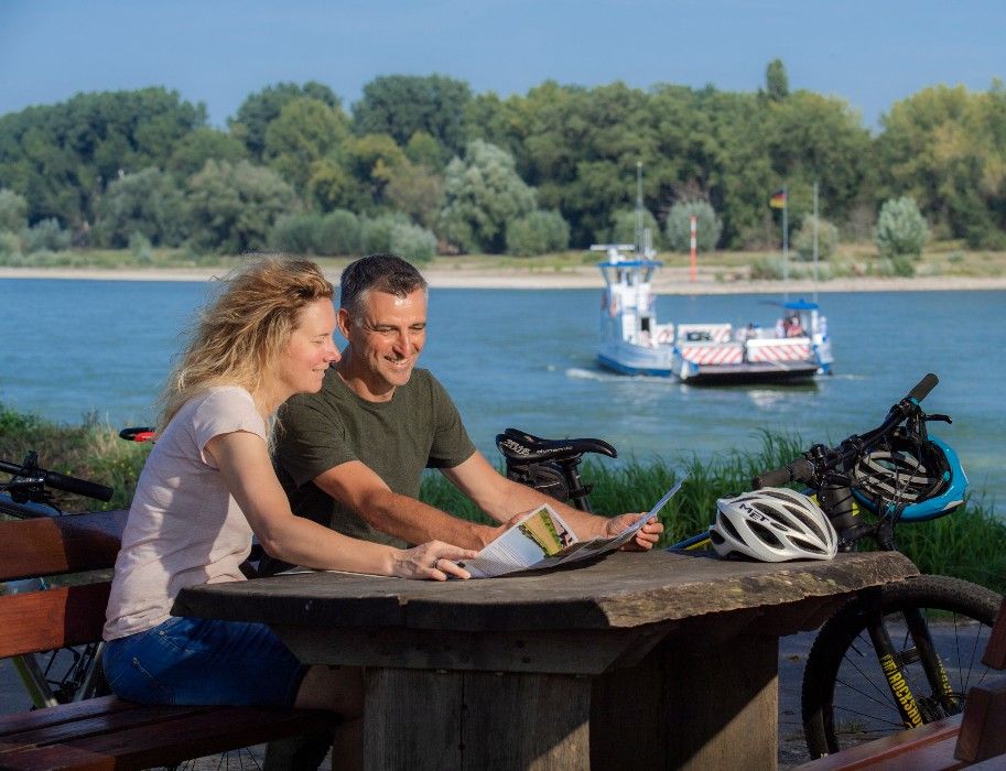 Ein Paar sitzt an einem Holztisch am Rhein in Zons, mit Fahrrädern und einer Fähre im Hintergrund.