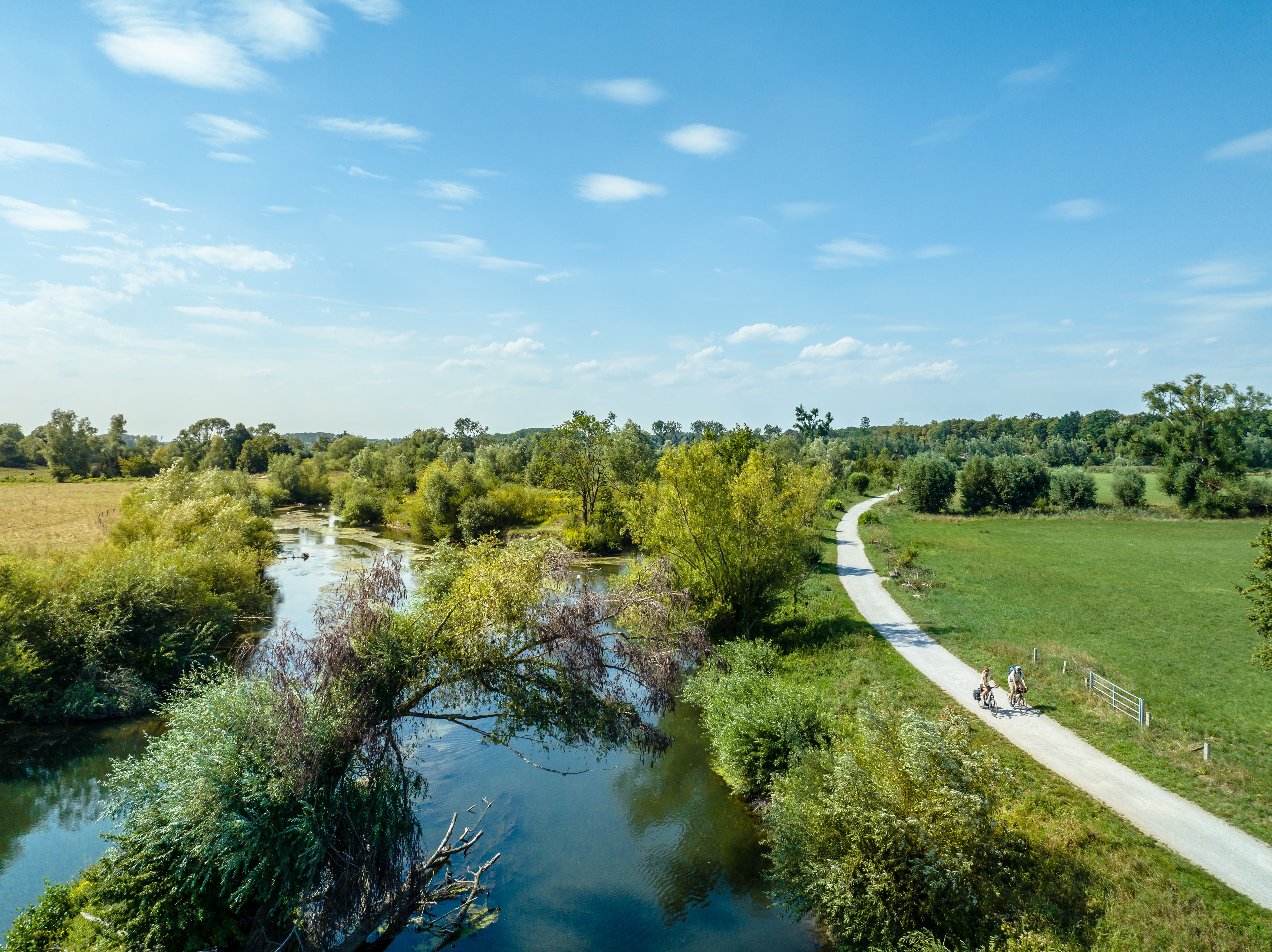 Landschaft mit Fluss, Bäumen und Radweg bei Lippstadt. Zwei Radfahrer sind auf dem Weg unterwegs. Blauer Himmel mit wenigen Wolken.