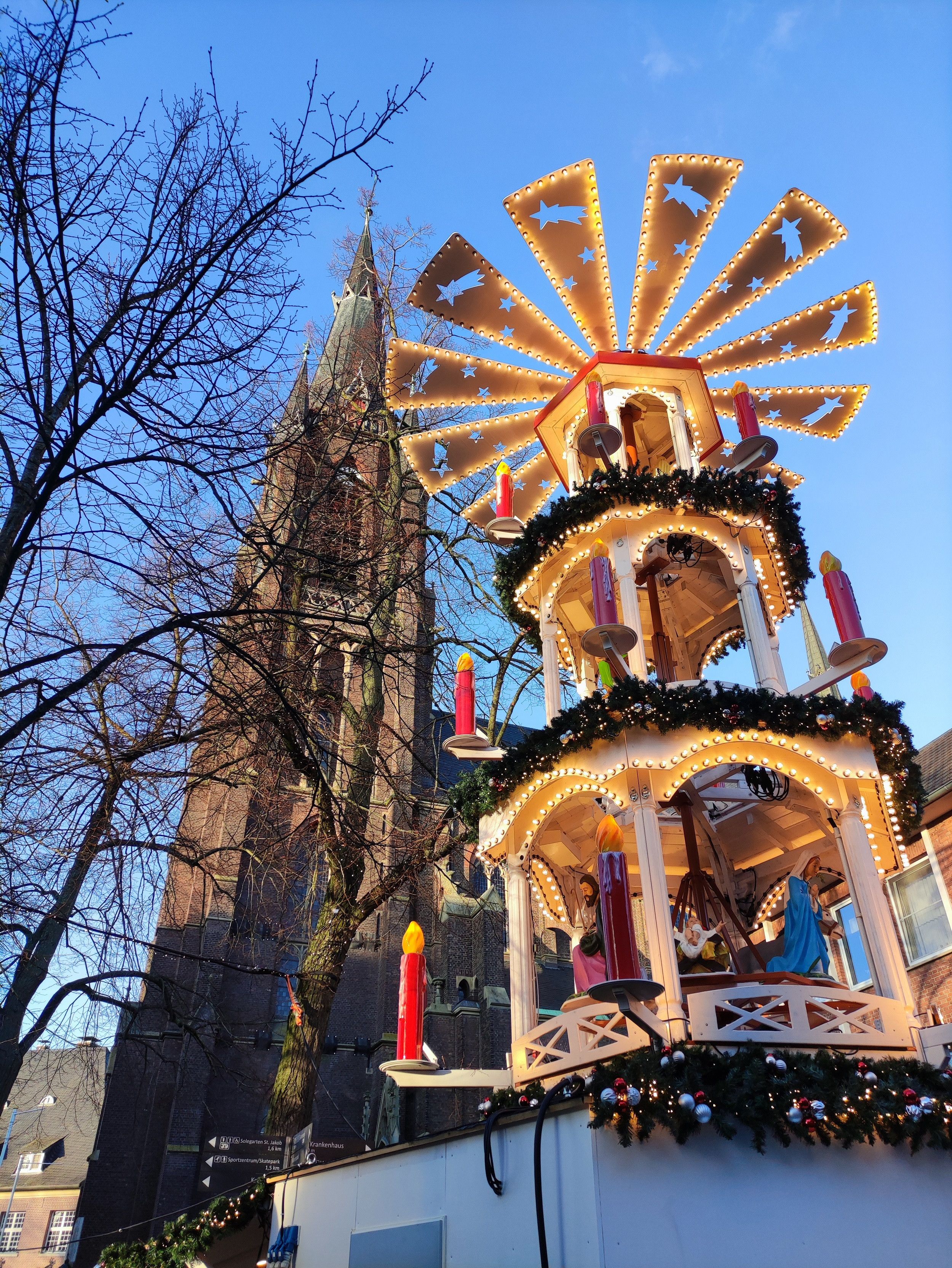 Eine beleuchtete Weihnachtspyramide mit Figuren steht vor einer hohen Kirchturmfassade in Kevelaer und läutet die Weihnachtsstimmung ein.