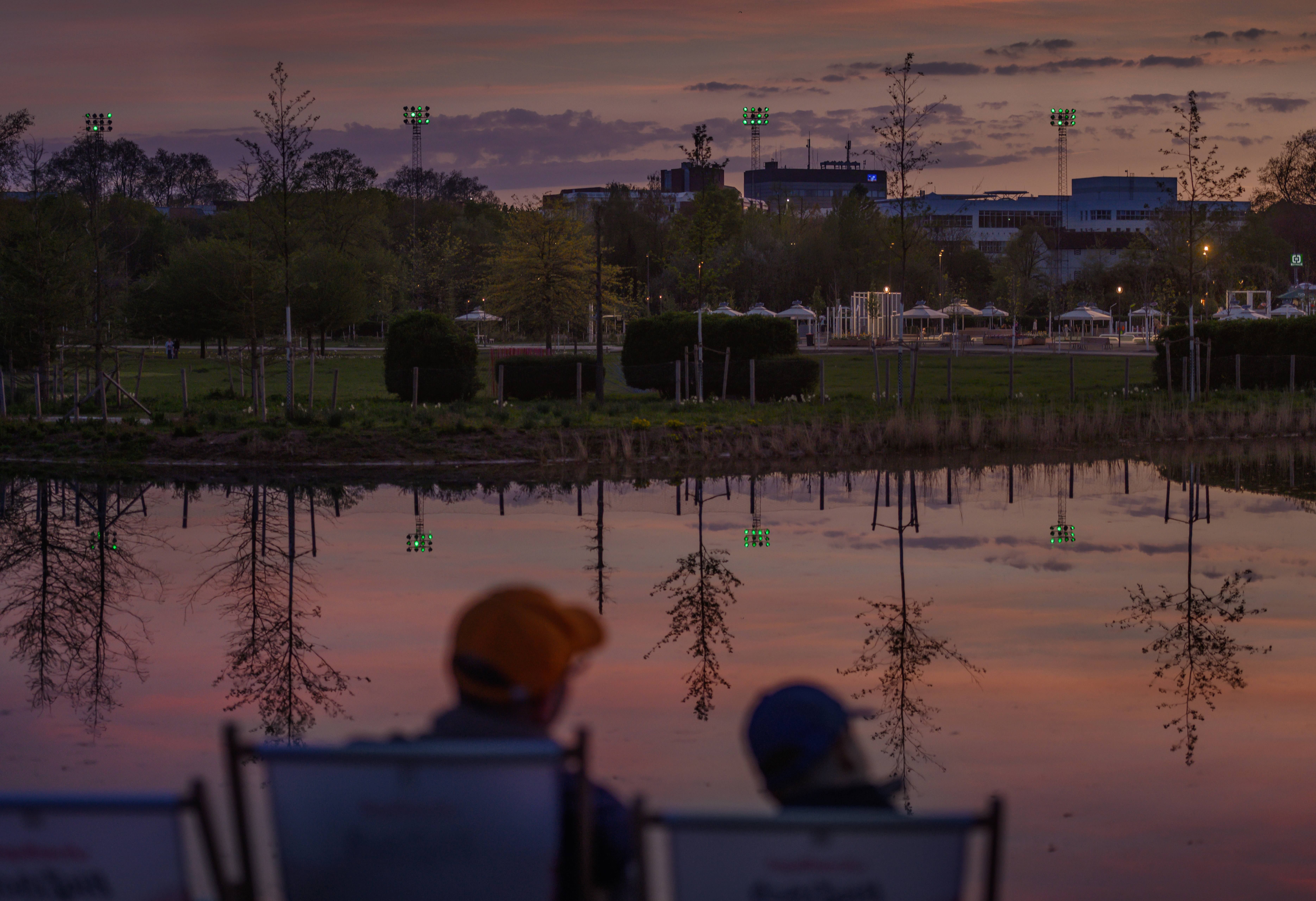Auf dem Gelände der Landesgartenschau Neuss 2026 sitzen zwei Personen in Liegestühlen vor einem See bei Sonnenuntergang.