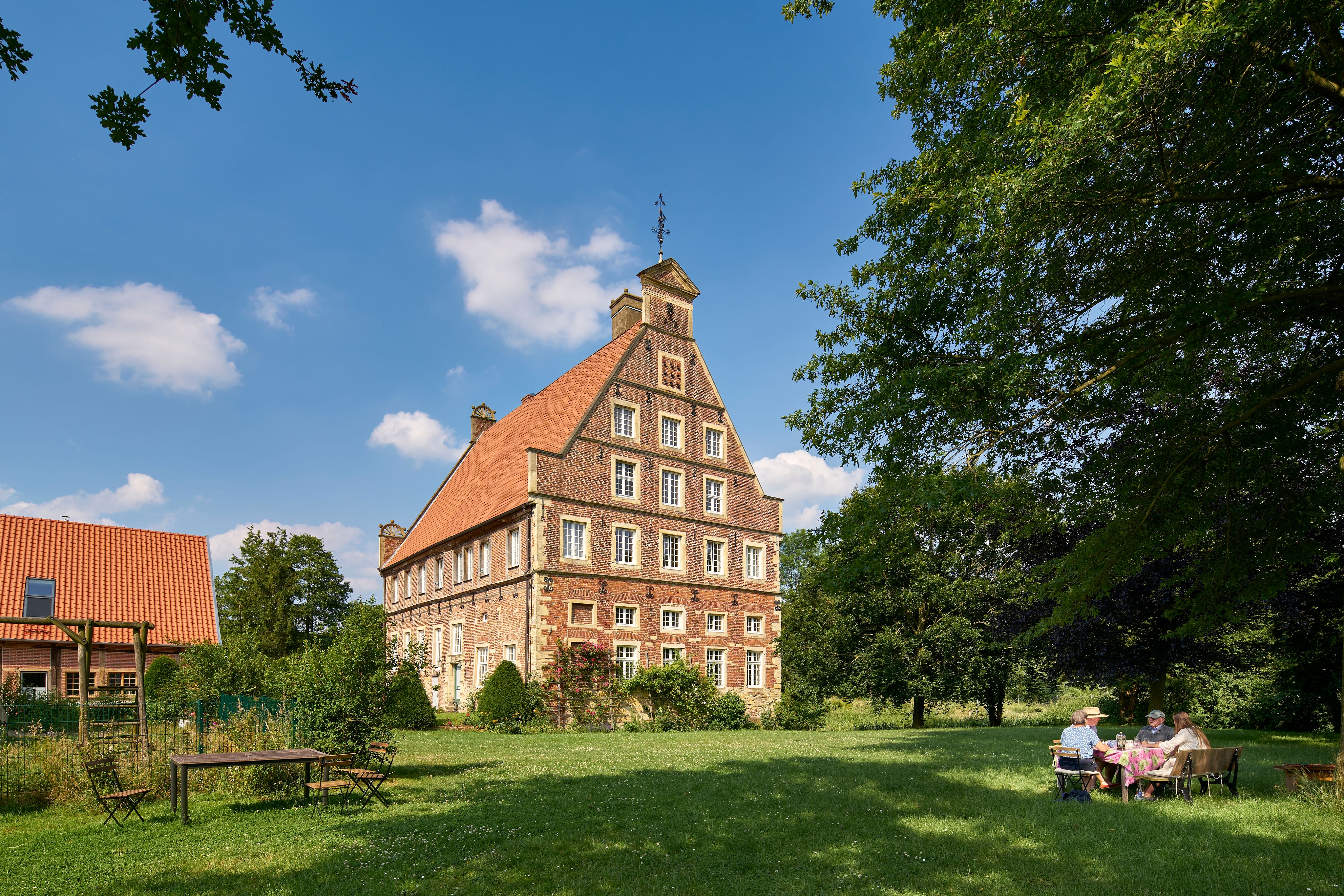 Haus Brückhausen. ein historisches Backsteingebäude mit rotem Dach in einer grünen Landschaft, Menschen sitzen im Garten.