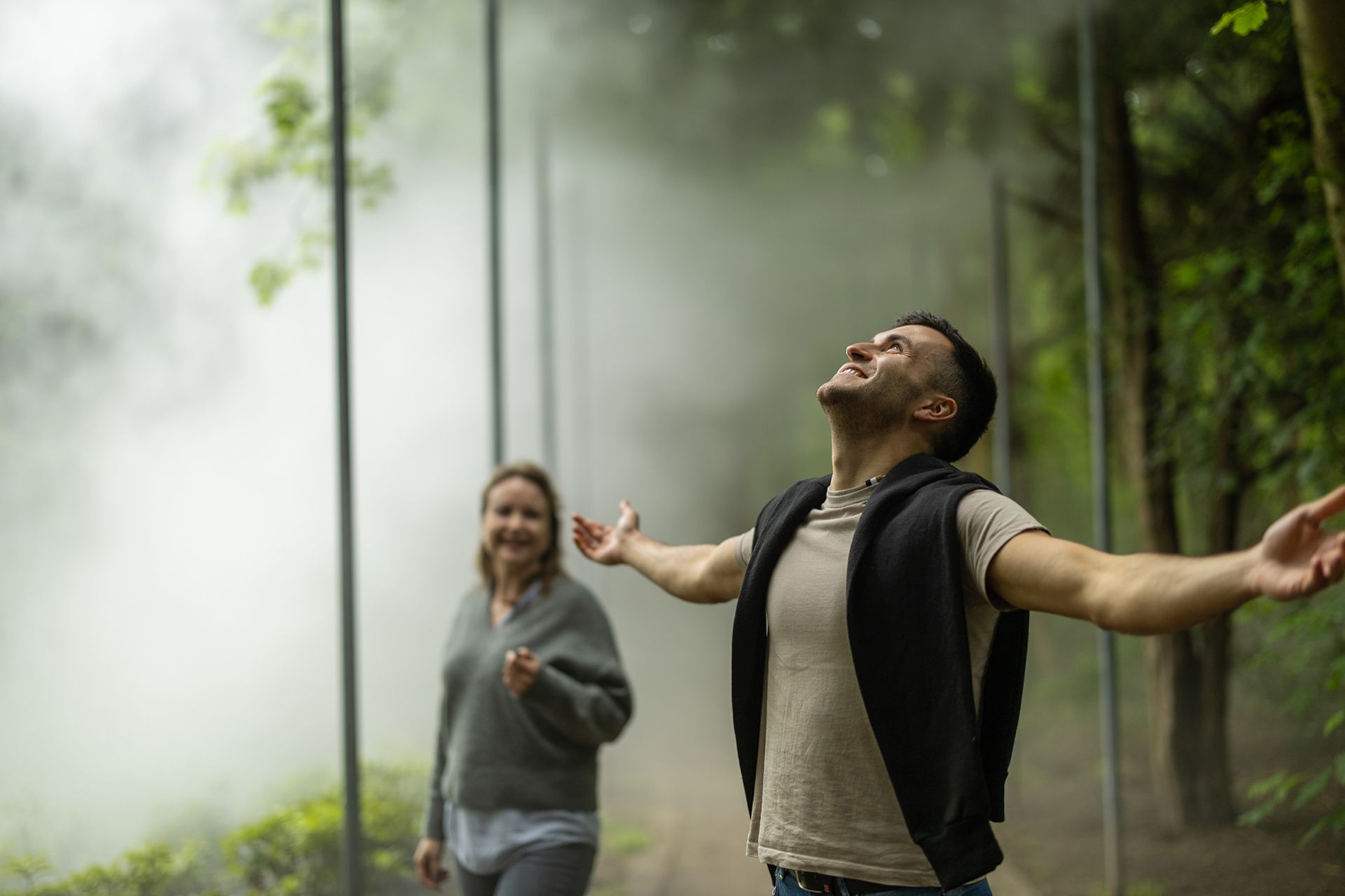 Ein Mann steht mit ausgebreiteten Armen in einem nebligen Wald, während eine Frau im Hintergrund lächelt.