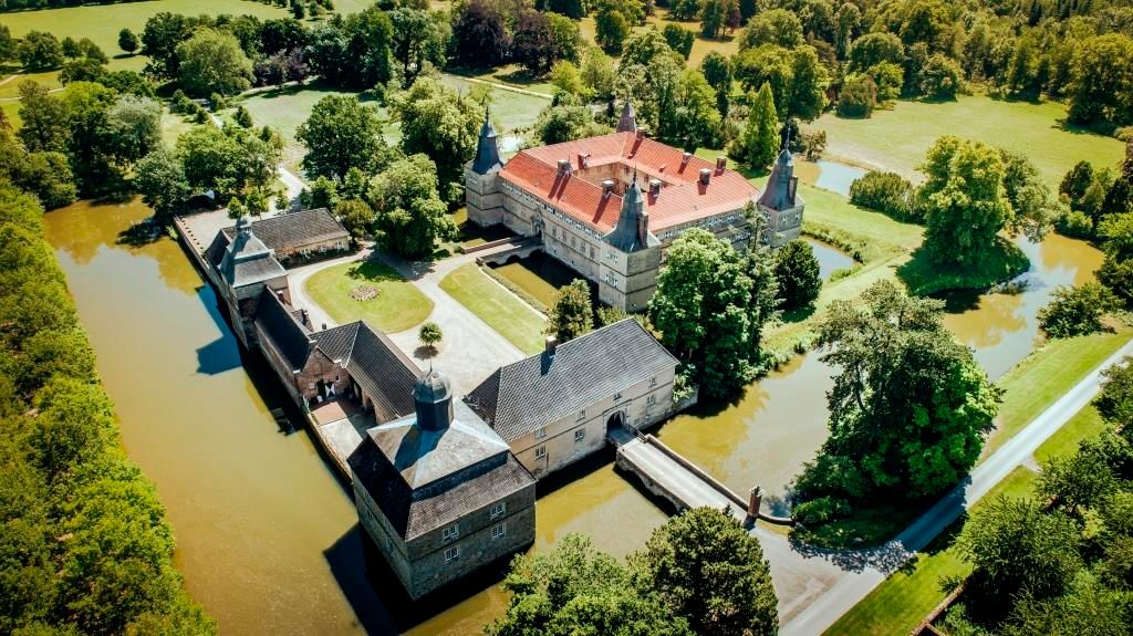 Luftaufnahme von Schloss Westerwinkel, umgeben von Wassergräben und grüner Landschaft. Das Schloss hat ein rotes Dach und ist von Bäumen umgeben.