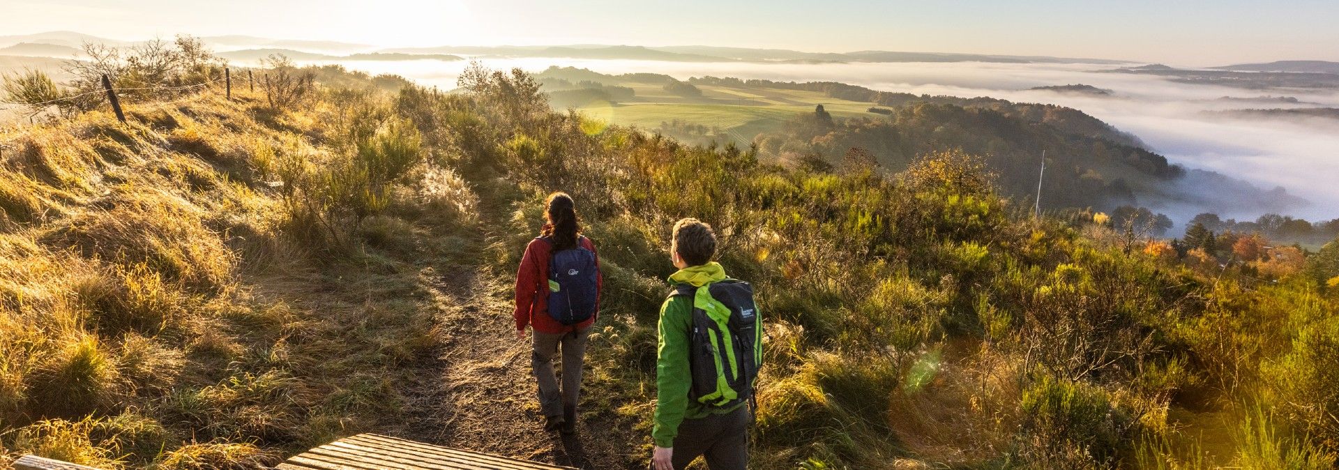 Zwei Wanderer auf einem Pfad in der Eifel mit Blick auf nebelverhangene Hügel im Sonnenaufgang.