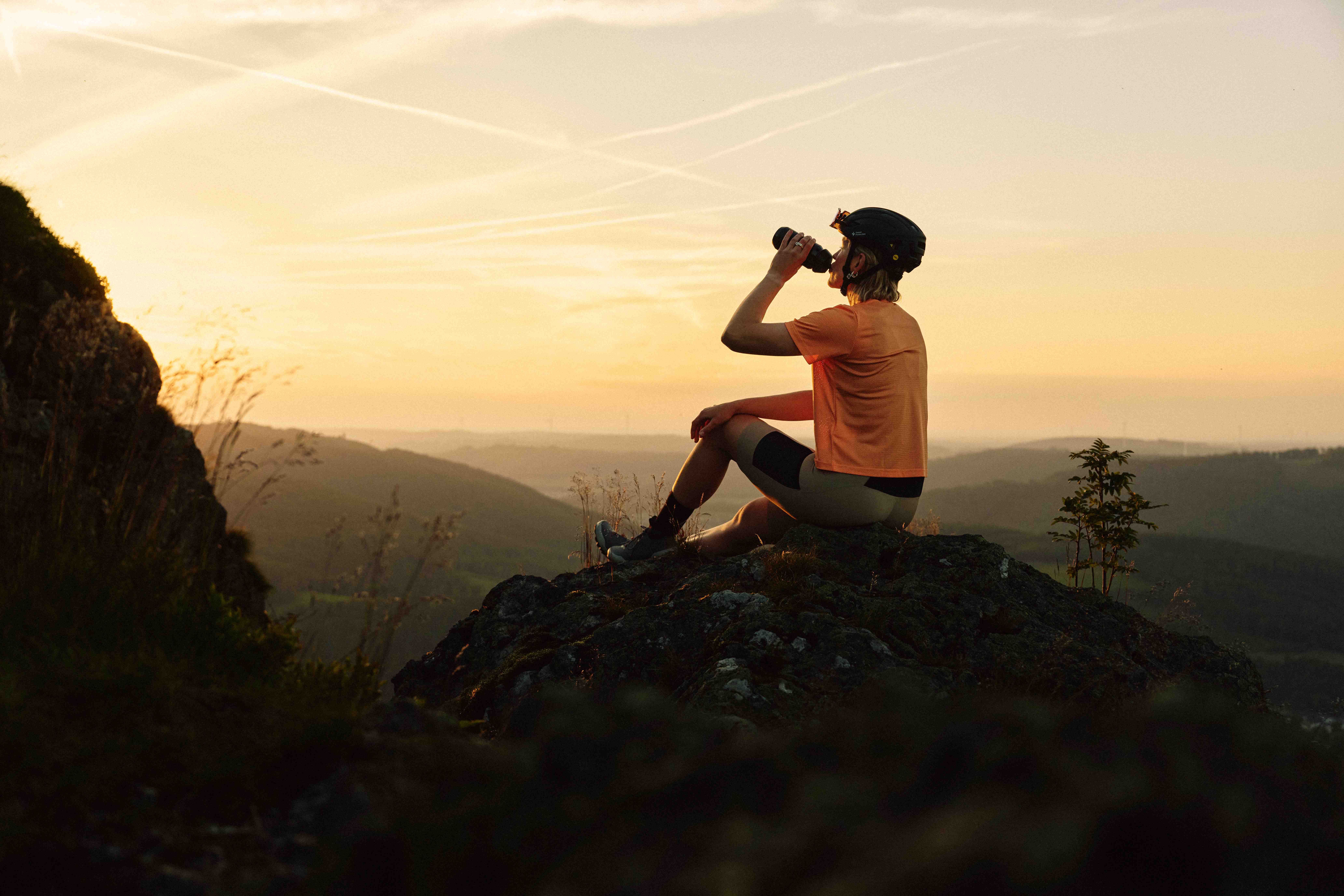 Eine Person mit Fahrradhelm sitzt auf einem Felsen und trinkt aus einer Flasche, während sie den Sonnenuntergang über einer hügeligen Landschaft betrachtet.