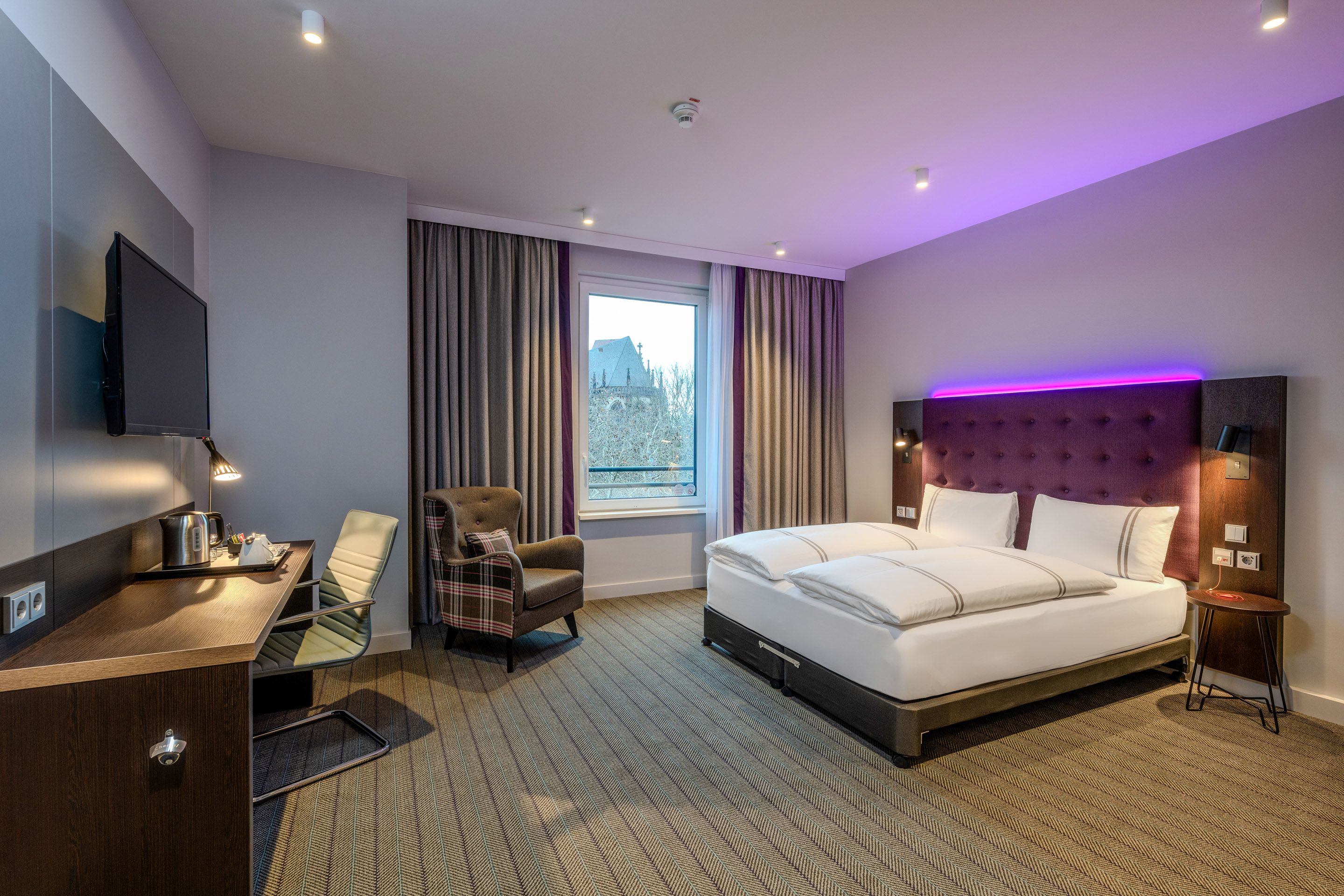 Modernes Hotelzimmer mit Doppelbett, Schreibtisch, Sessel und Fenster mit Blick nach draußen.