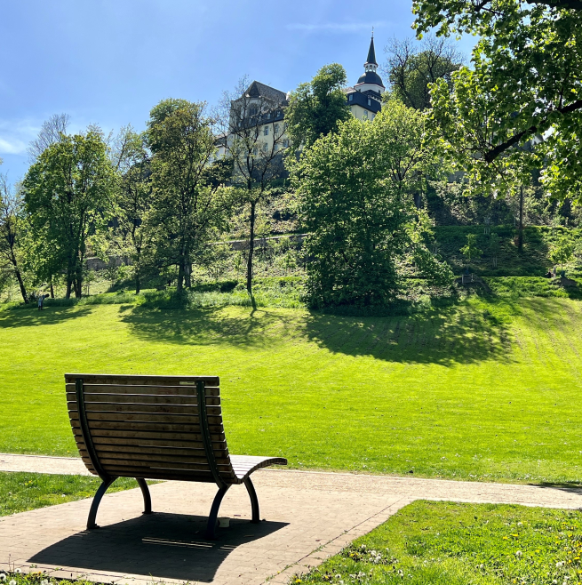 Bank im Park mit Blick auf das Schloss Siegburg auf dem Hügel, umgeben von Bäumen und sonniger Wiese.