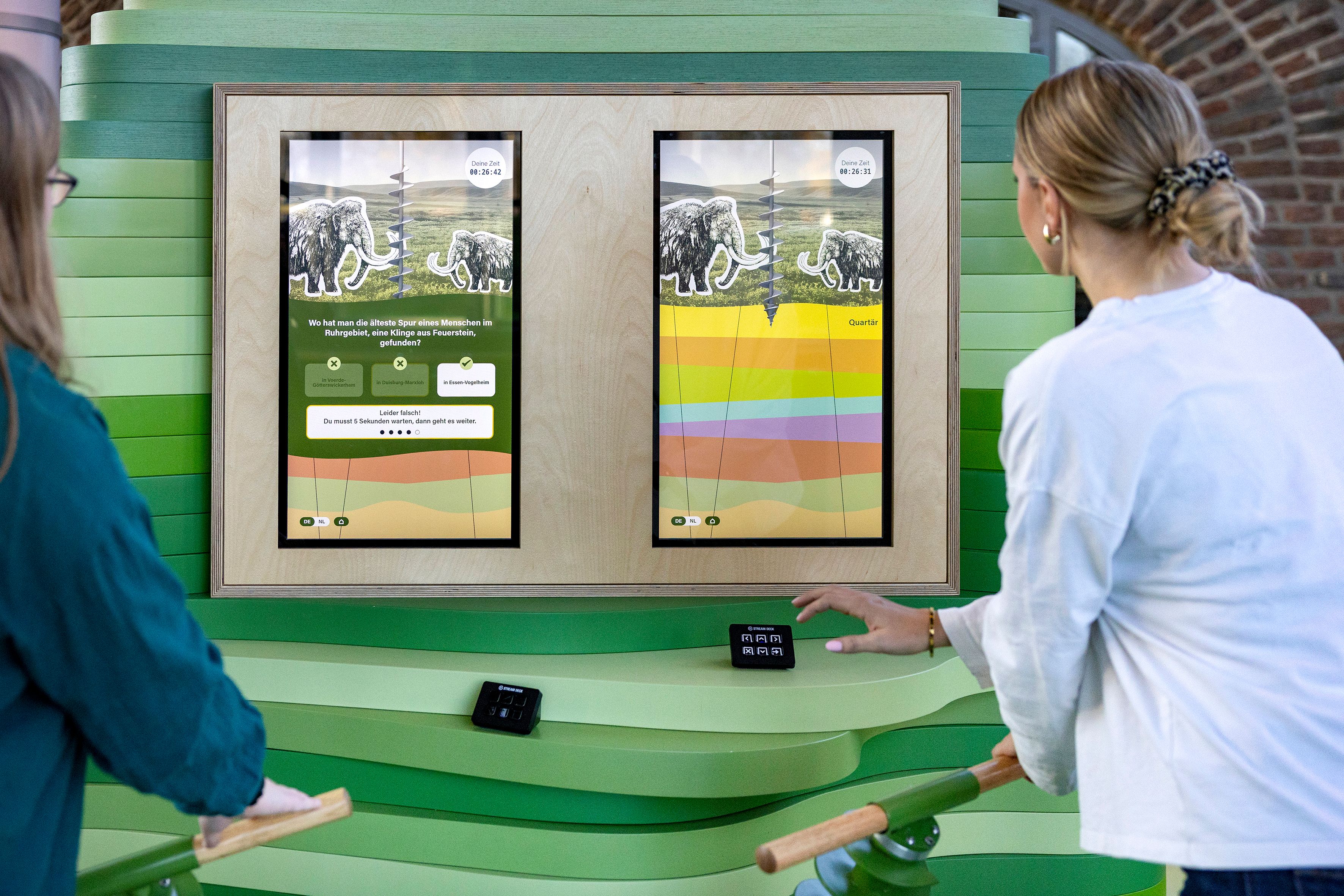 Zwei Personen interagieren mit einem digitalen Display in einem GeoPark. Das Display zeigt Informationen über Erdgeschichte und Mammuts.