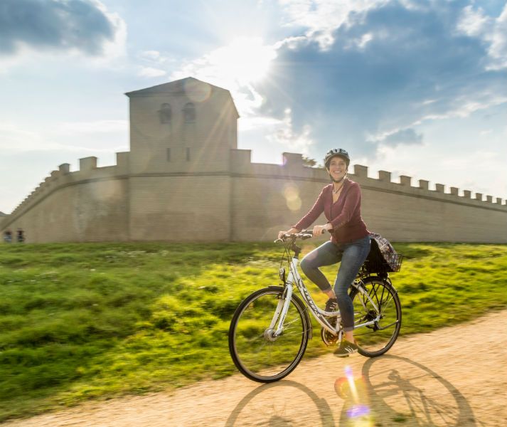 Eine Person fährt bei Sonnenschein auf einem Fahrrad vor einer römischen Mauer entlang. Der Himmel ist teils bewölkt