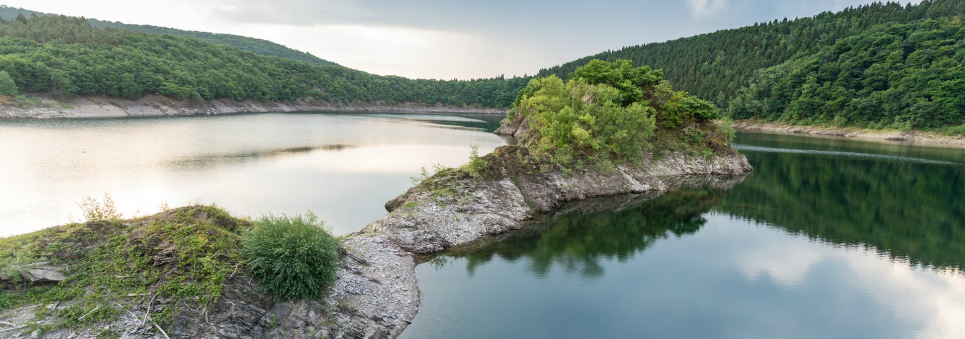 Die Urfttalsperre mit ruhigem Wasser, umgeben von bewaldeten Hügeln und einer kleinen, bewachsenen Landzunge im Vordergrund.