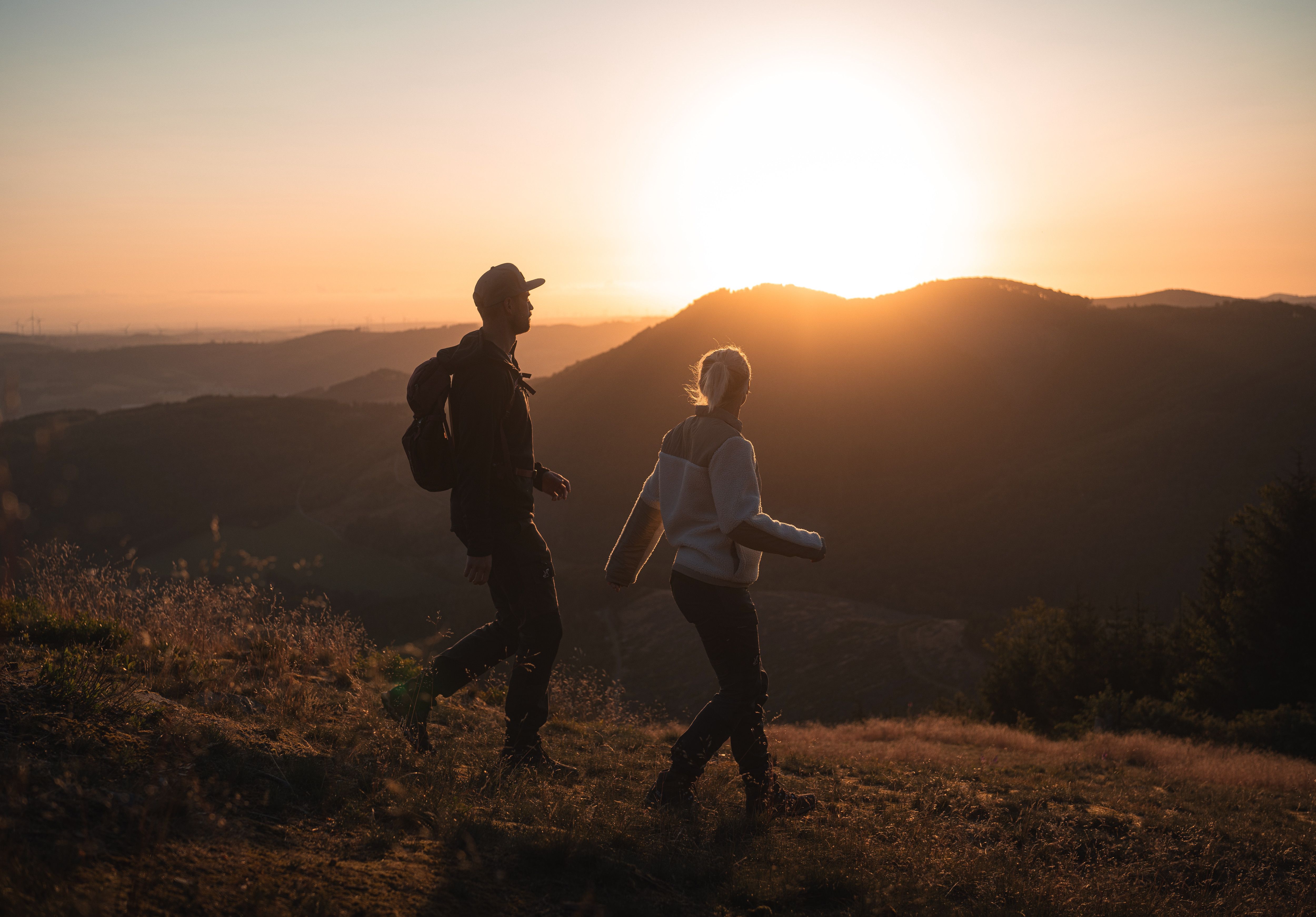 Zwei Wandernde bewegen sich im warmen Licht des Sonnenuntergangs über einen Höhenweg. Die umliegenden Hügel zeichnen sich als sanfte Silhouetten am Horizont ab und verleihen der Szene eine stimmungsvolle, naturnahe Atmosphäre.