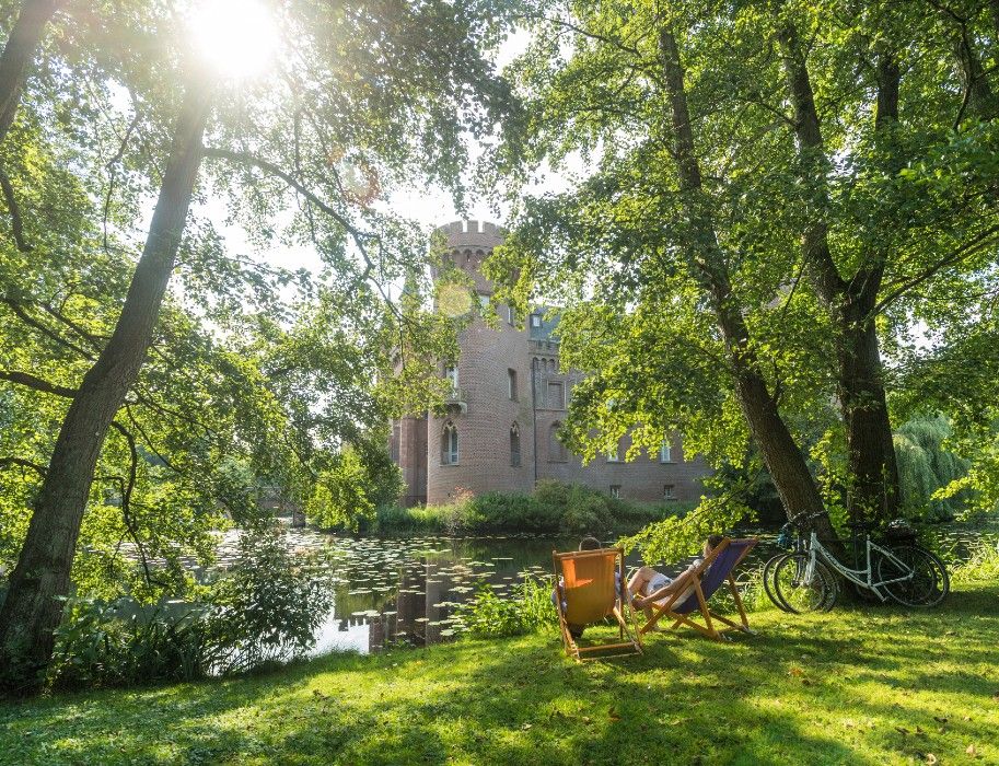 Schloss Moyland im Sonnenlicht, umgeben von Bäumen und einem Teich. Zwei Liegestühle laden zum Verweilen ein
